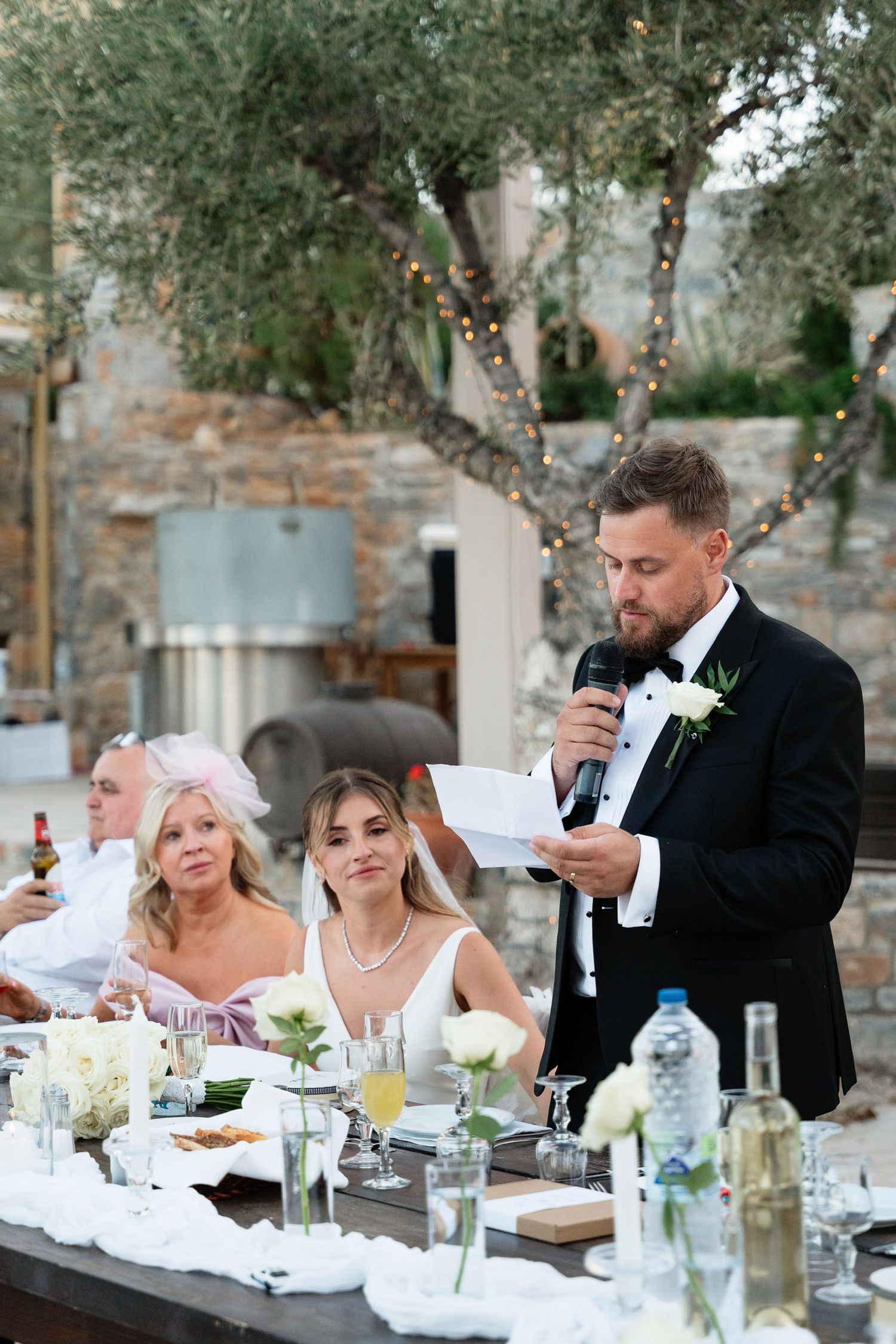 A wedding reception dinner outdoors with guests seated at a table while someone gives a speech at sunset.
