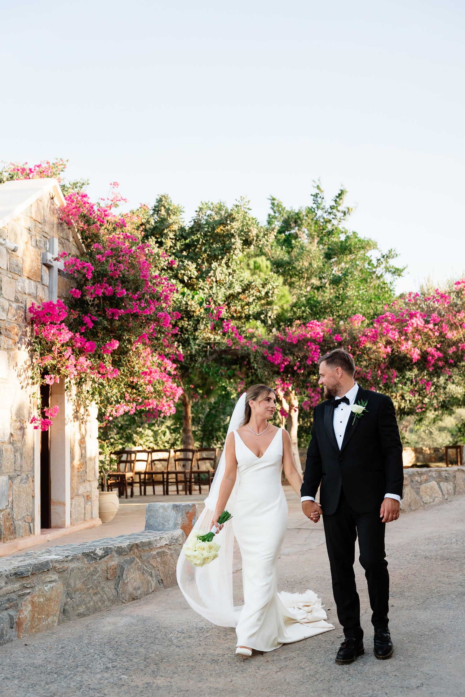 A couple in wedding attire walks along a stone path lined with vibrant pink bougainvillea flowers in a garden setting.