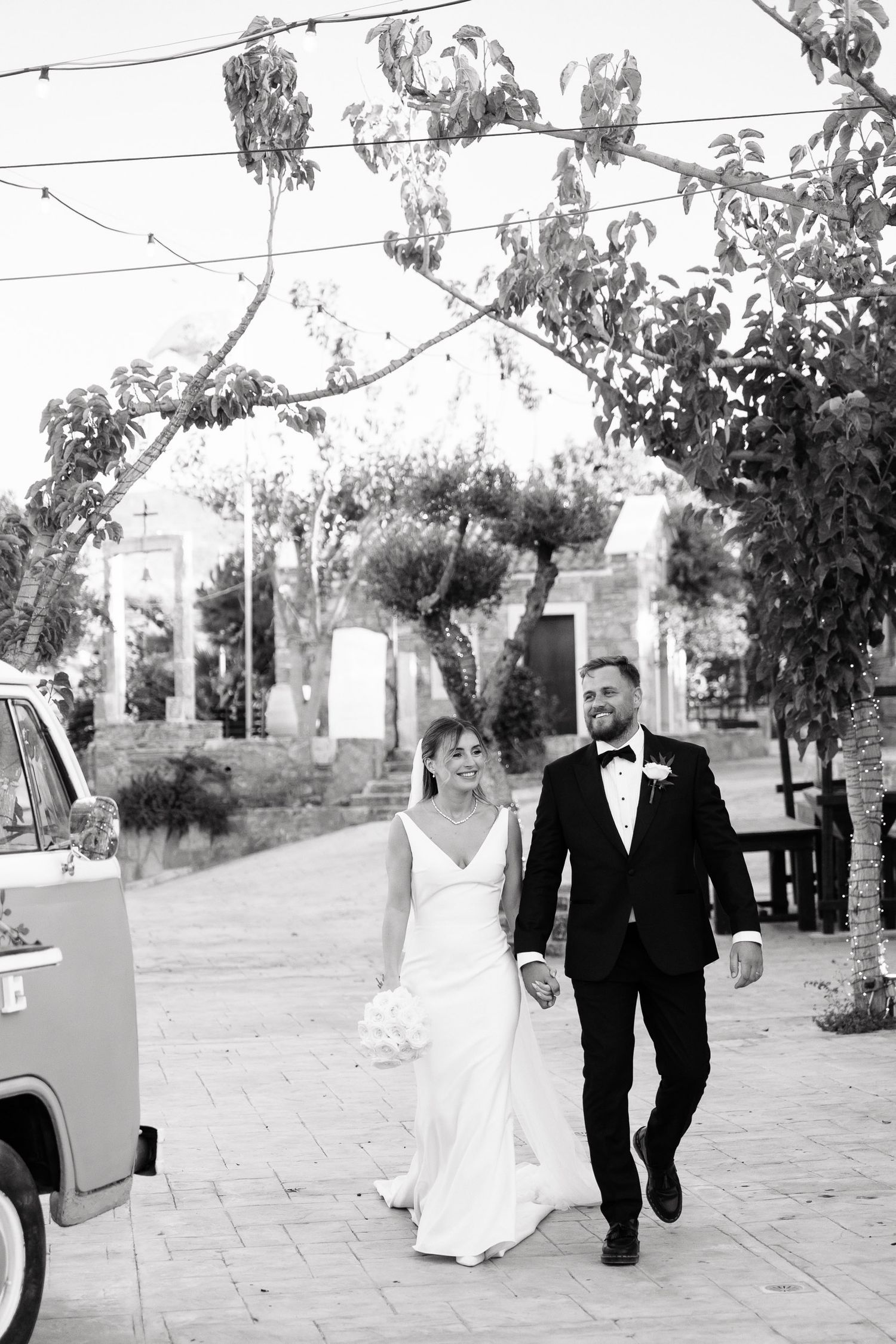 A couple in wedding attire walking together on a path with trees and power lines visible in the black and white photo.
