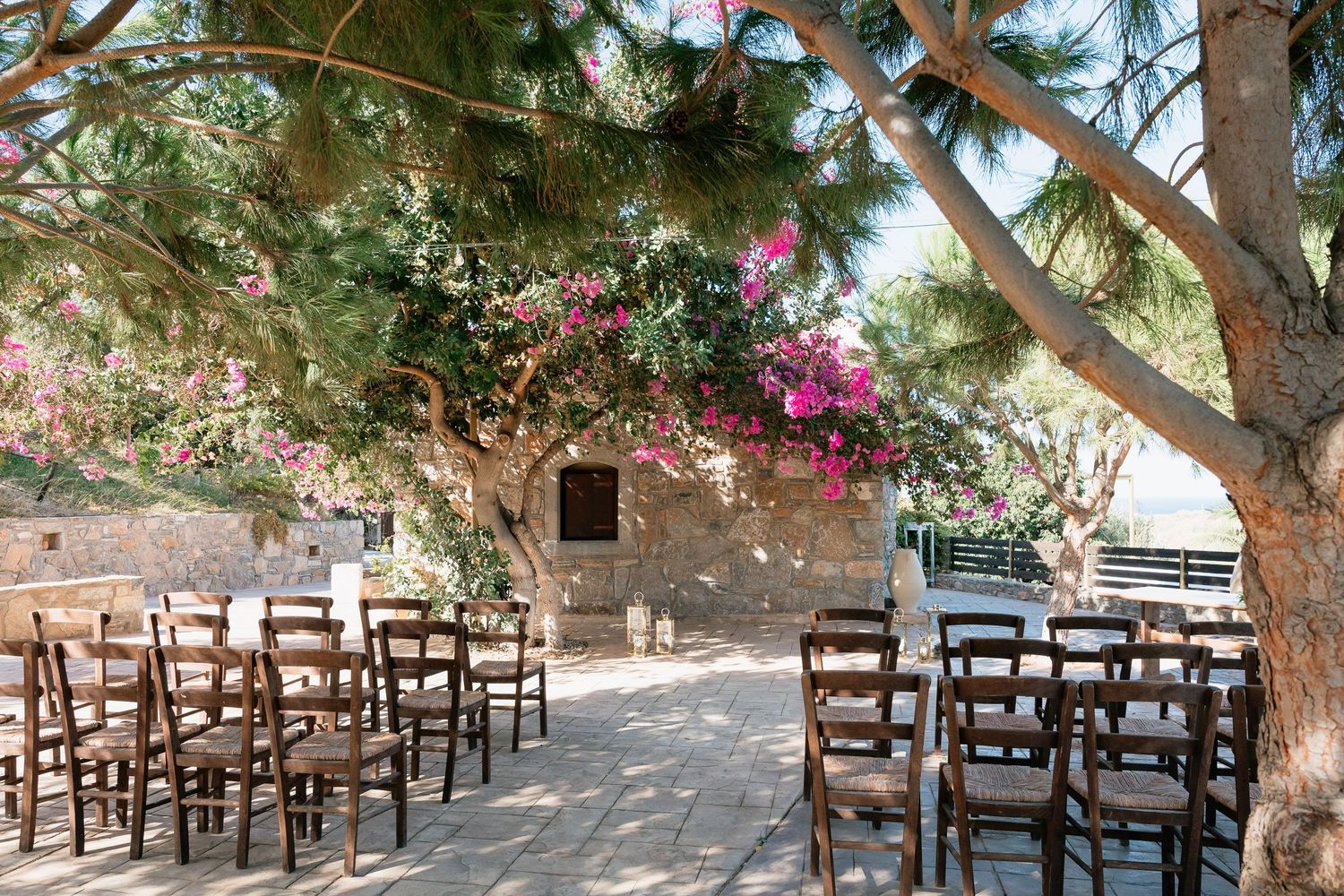 Rustic wooden chairs line a shaded outdoor terrace with stone walls and dappled sunlight filtering through trees.