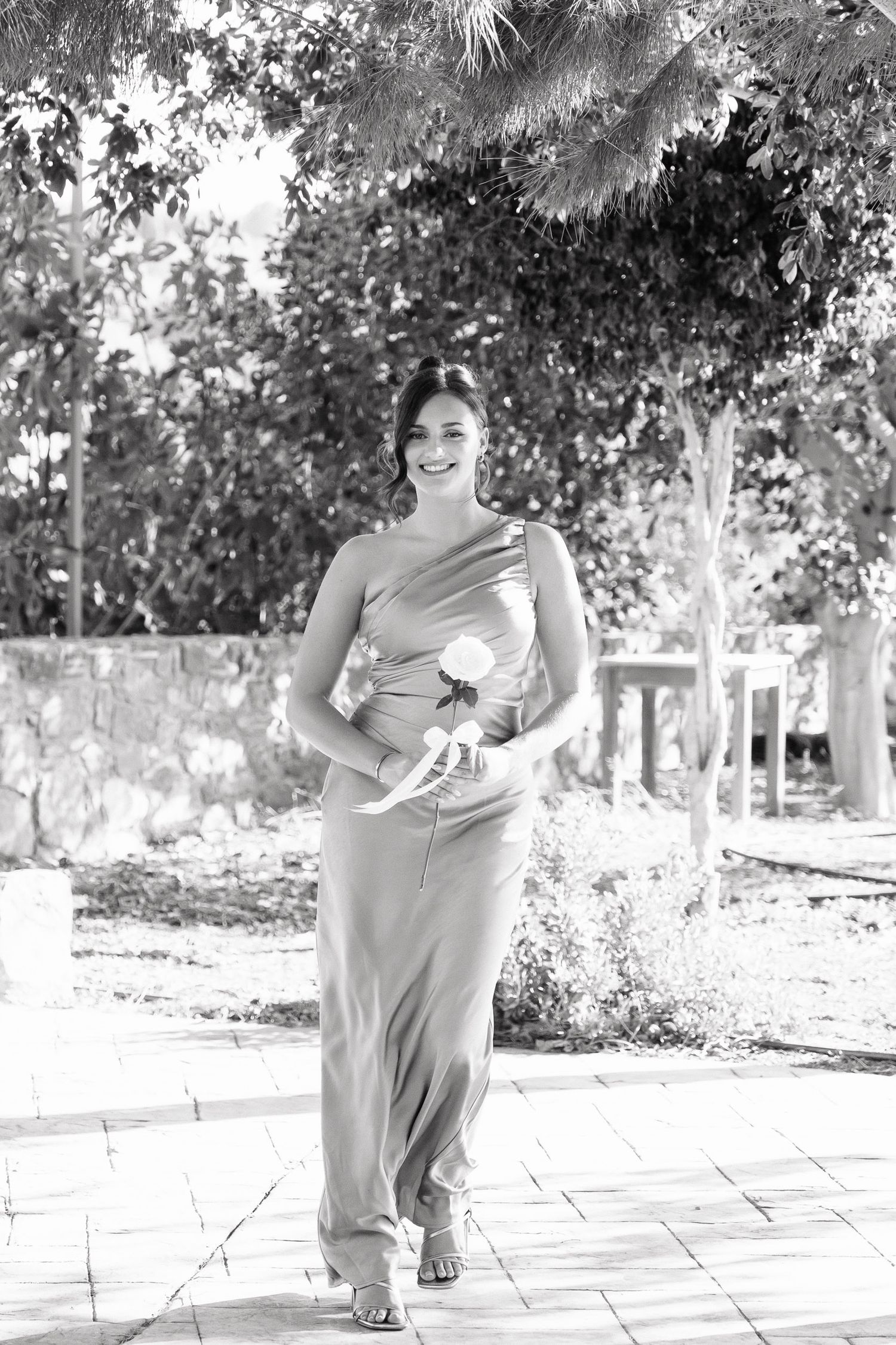 A person in light clothing dances joyfully in a garden setting with trees and paved walkways in the background.