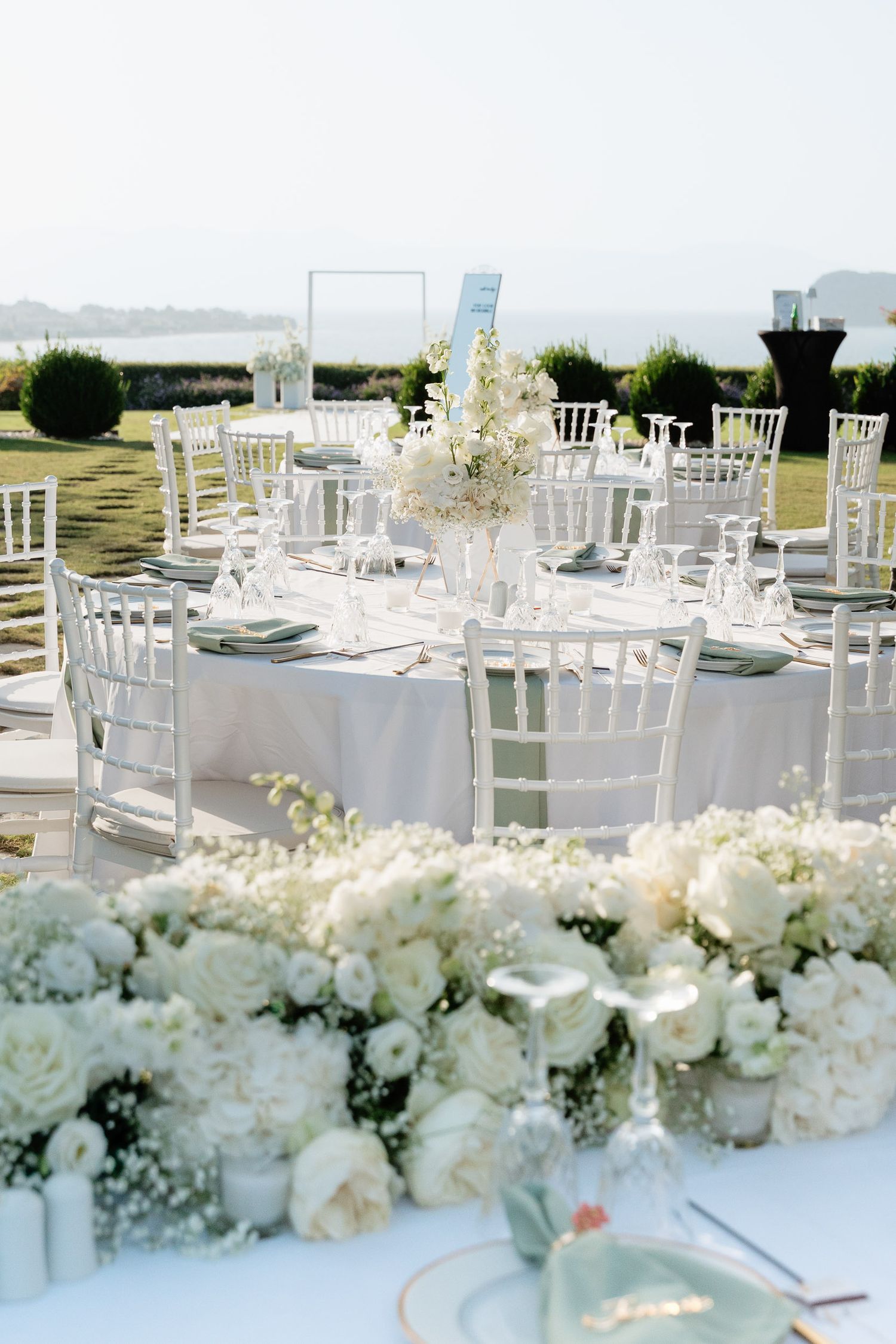 White Chiavari chairs and tables set for an outdoor wedding reception with lush white floral centerpieces.