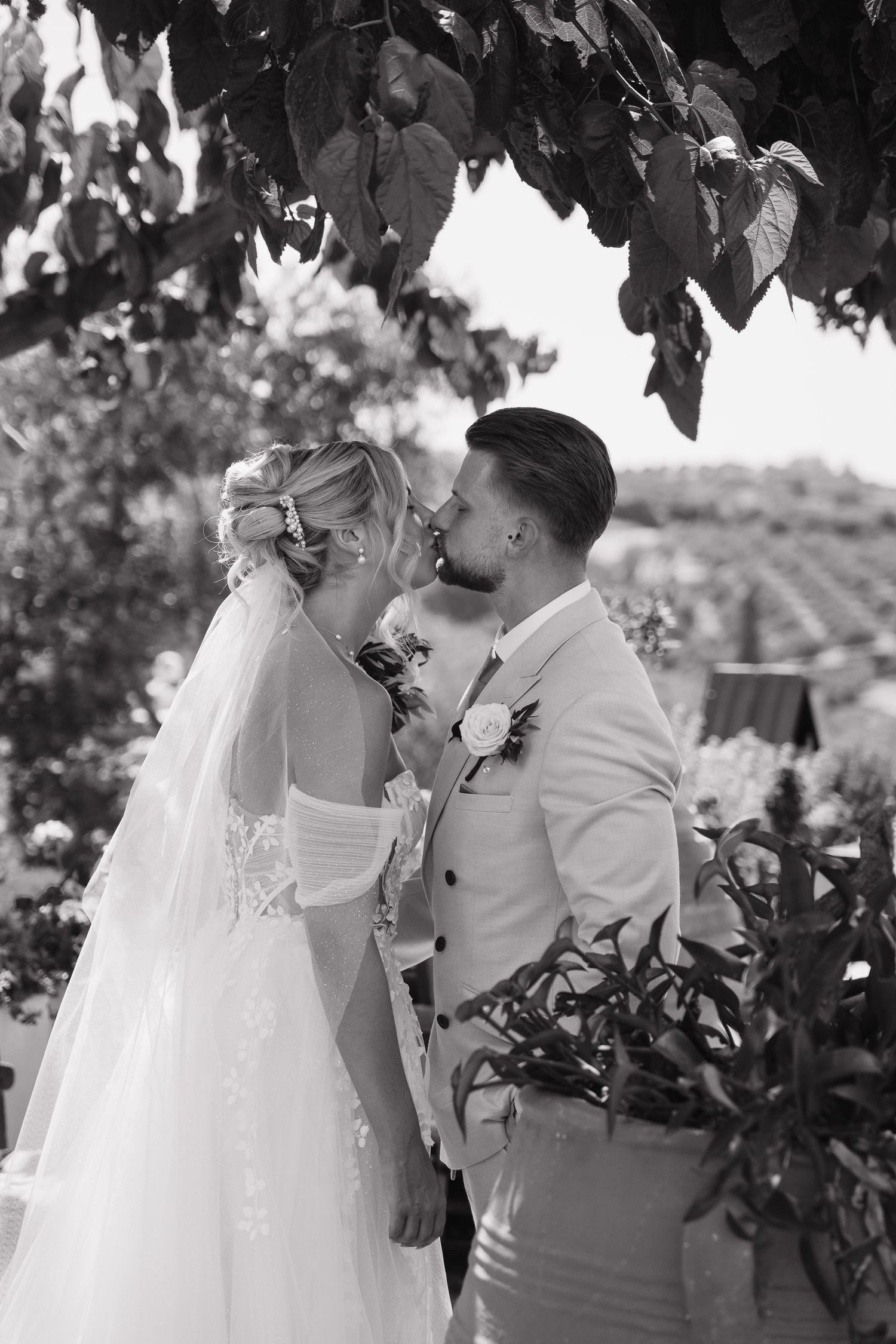 A romantic black and white wedding photograph captured under tree leaves showing a tender moment between newlyweds.