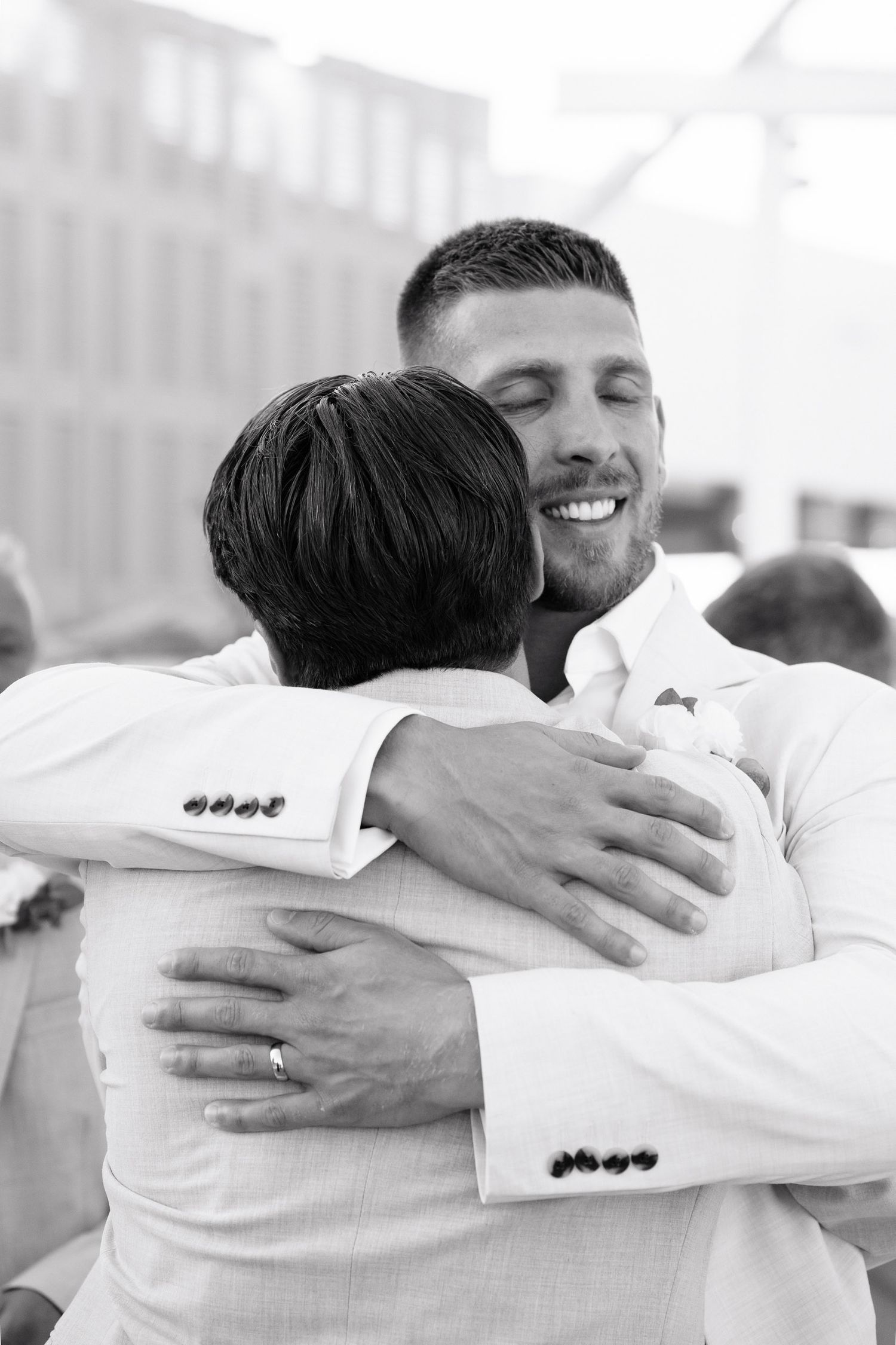 Two people in white clothing embrace in a warm hug at what appears to be a wedding celebration.