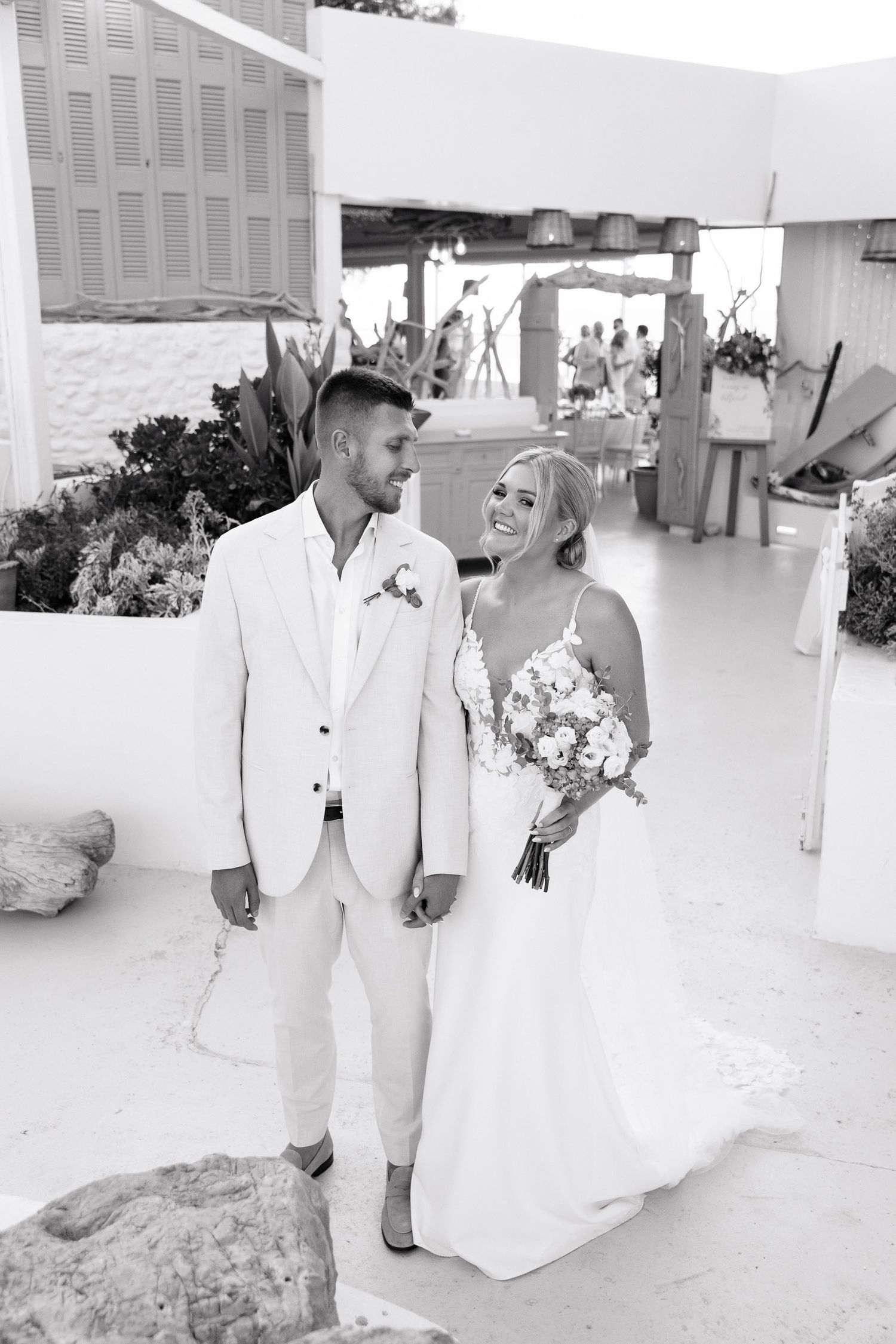 A black and white wedding photograph showing a couple in matching white attire sharing a joyful moment outdoors.