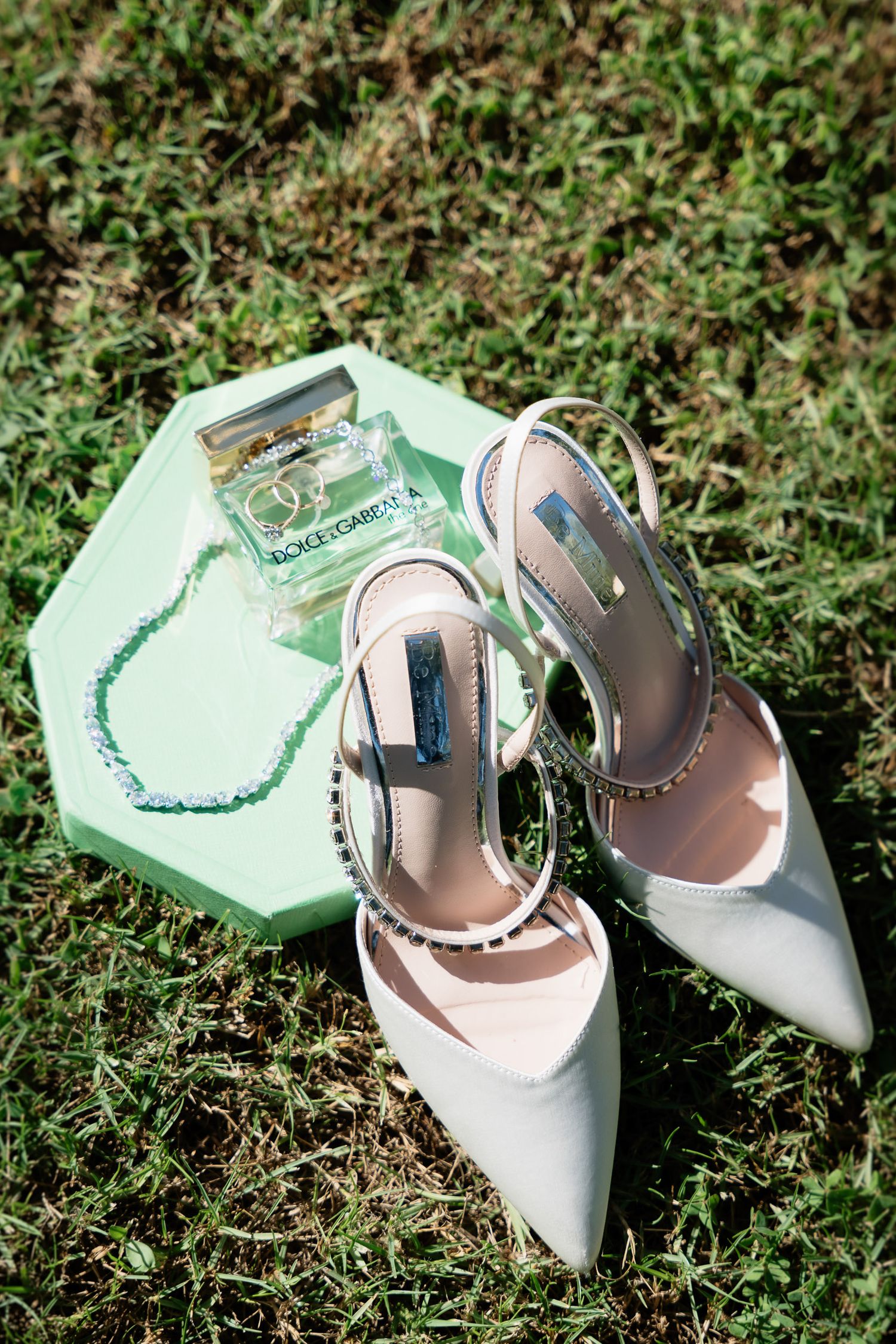 Elegant grey pointed-toe slingback heels with pearl details sit beside a mint green clutch on grass.