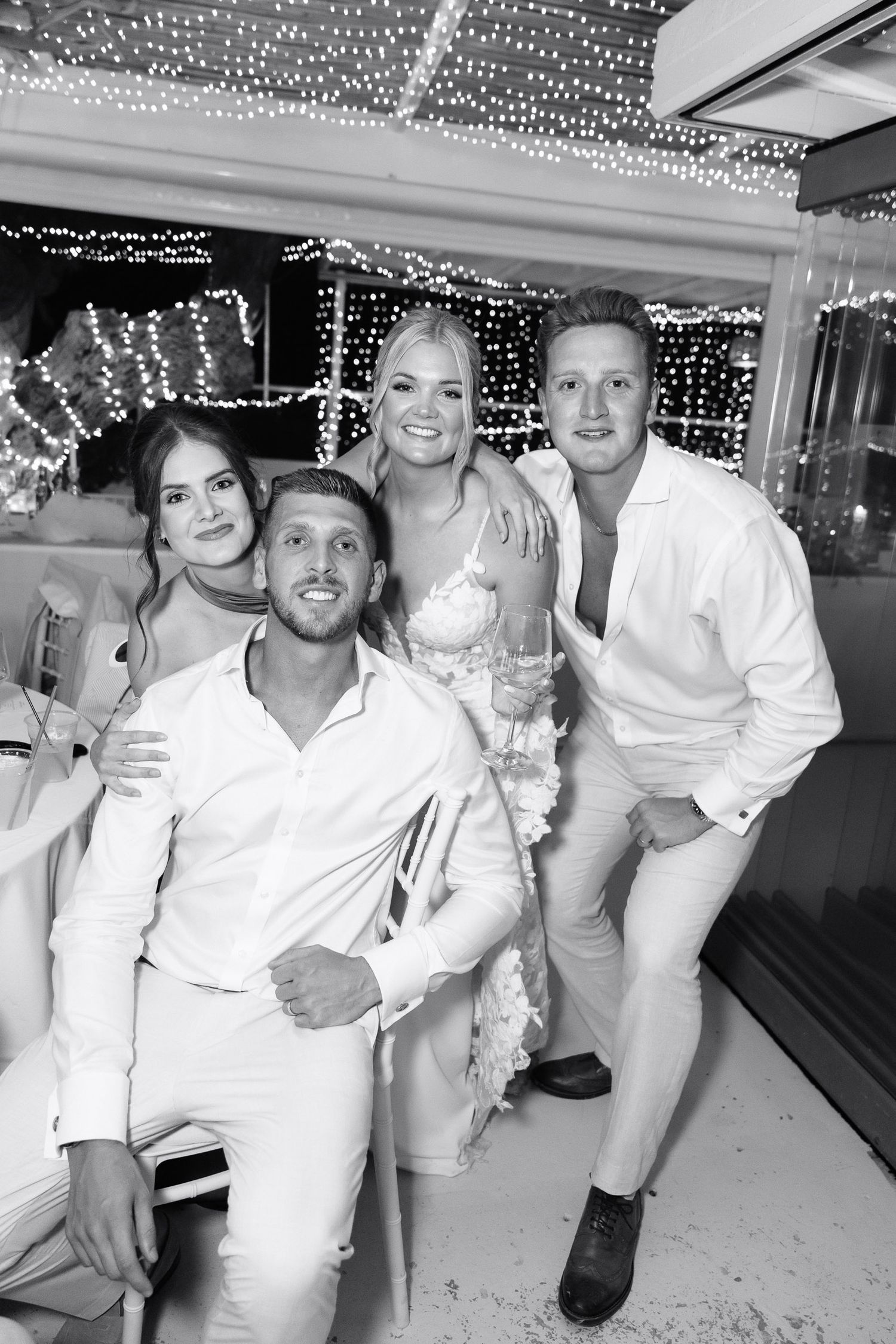 Black and white photo of a wedding celebration with people posing together under decorative string lights.