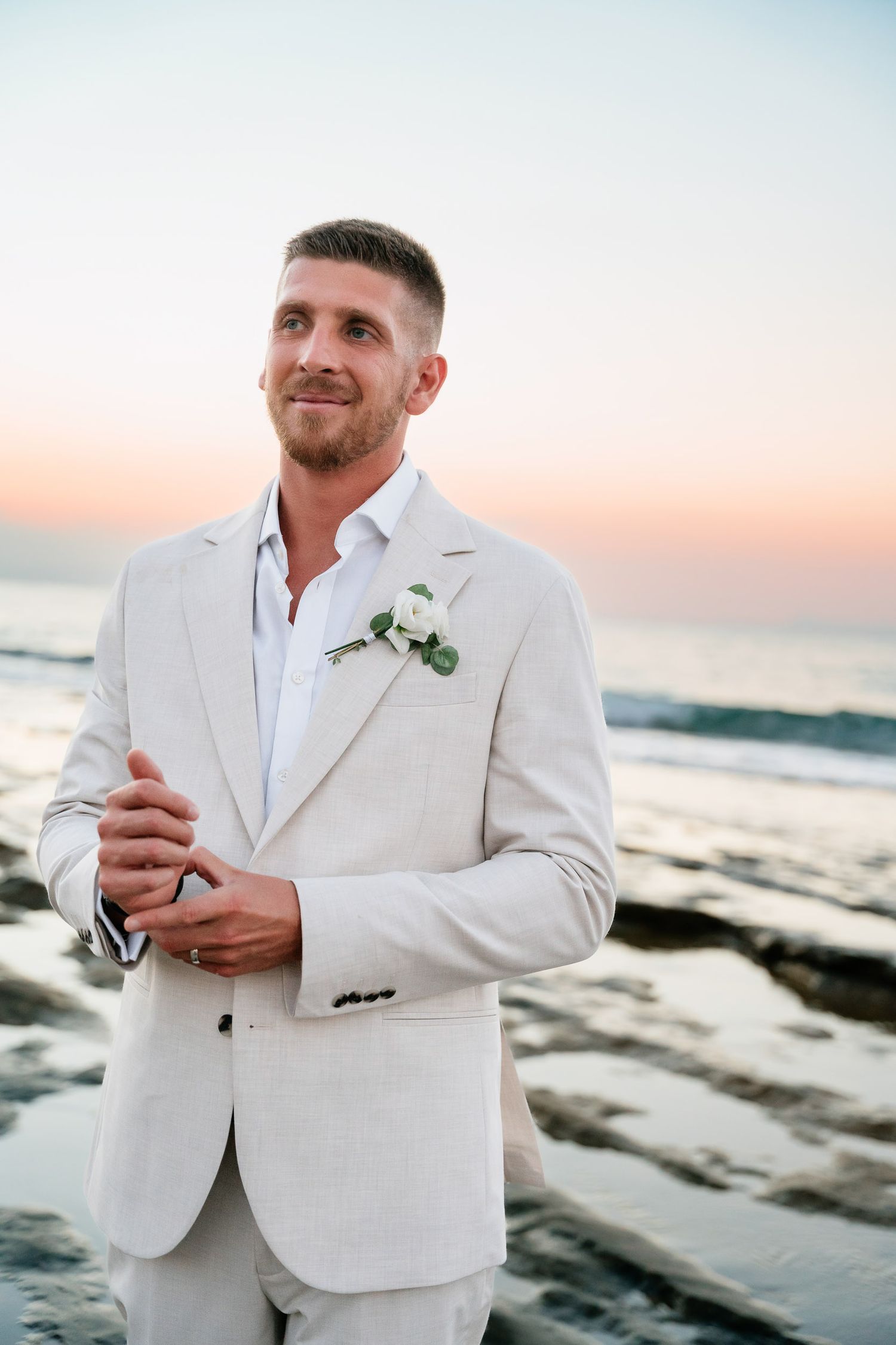 A person in a white suit jacket poses on a rocky beach during sunset.