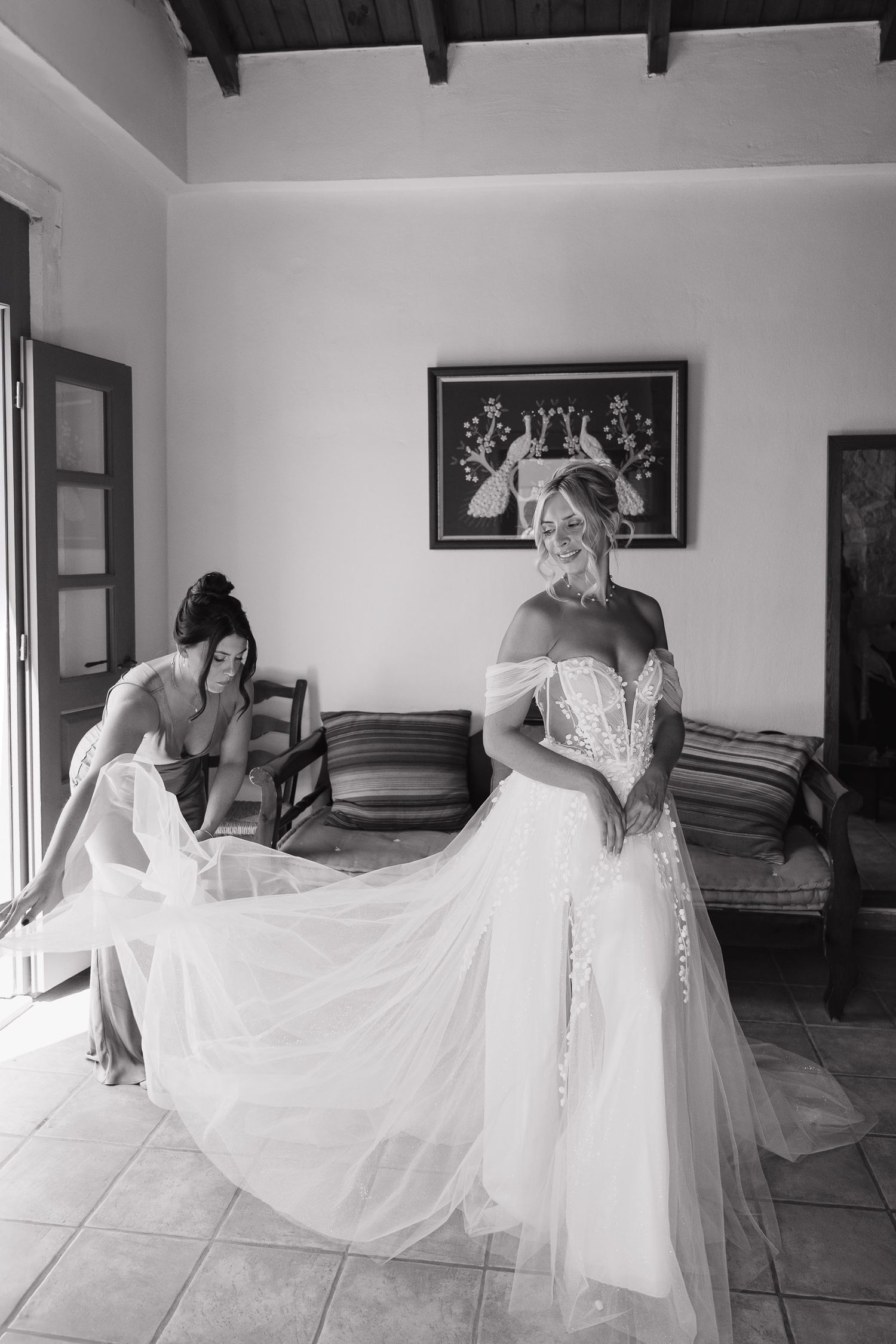 A black and white photo of a bride in an elegant off-shoulder wedding gown having her dress adjusted before the ceremony.