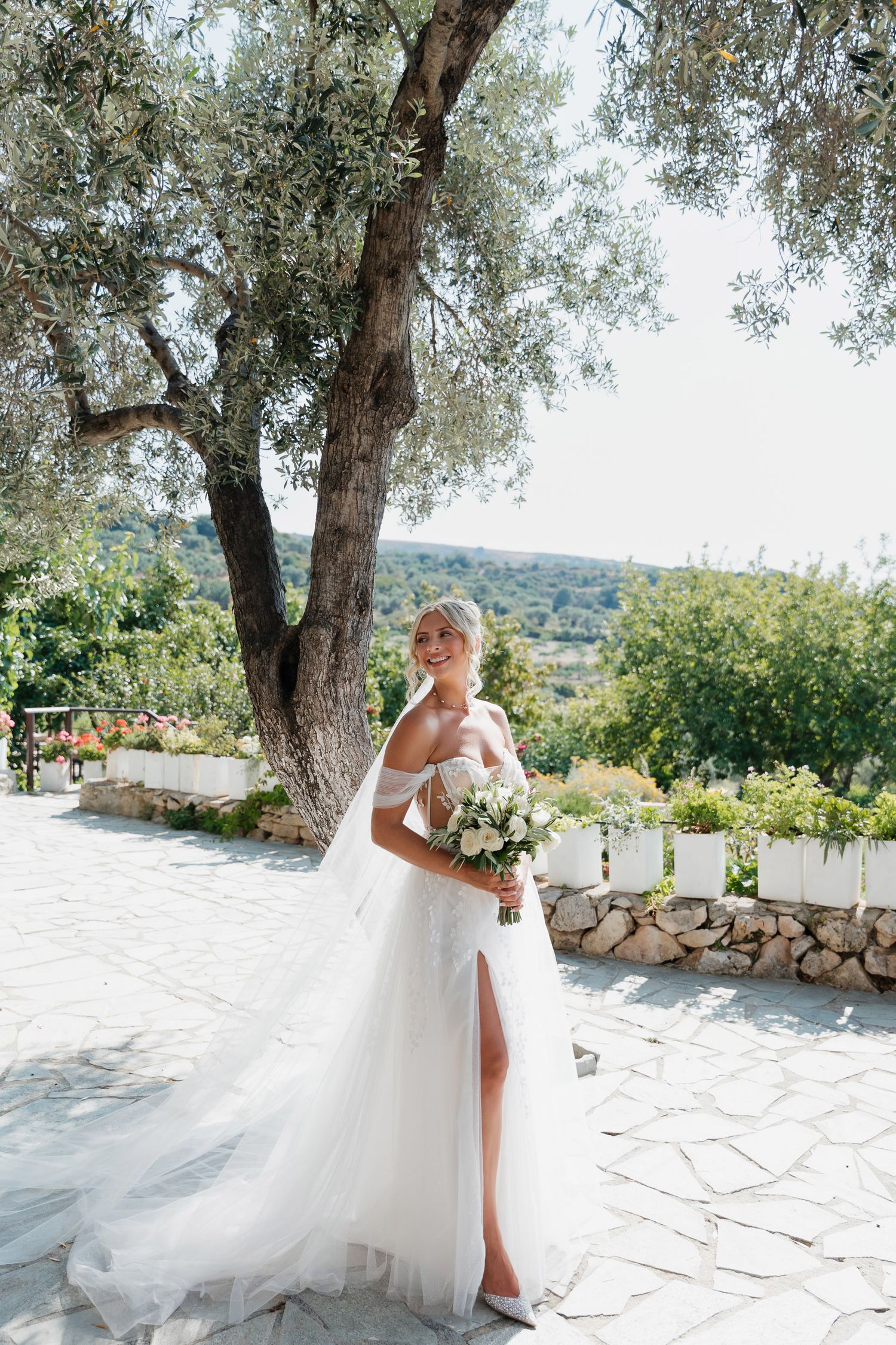 A flowing white wedding dress with a thigh slit is showcased in an outdoor Mediterranean setting with olive trees.