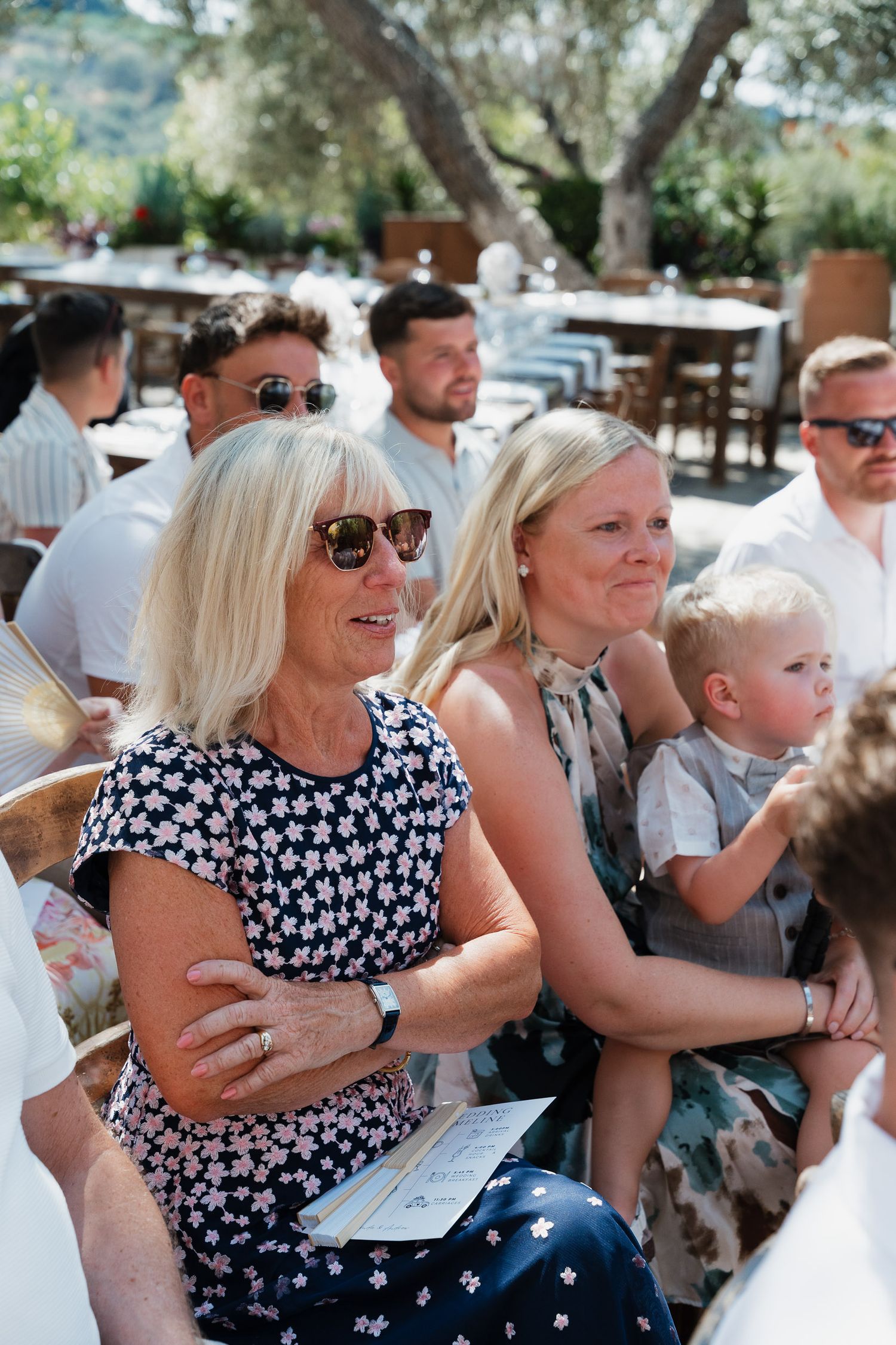 A group of people sitting outdoors at a sunny venue, wearing sunglasses and summer attire while watching an event.
