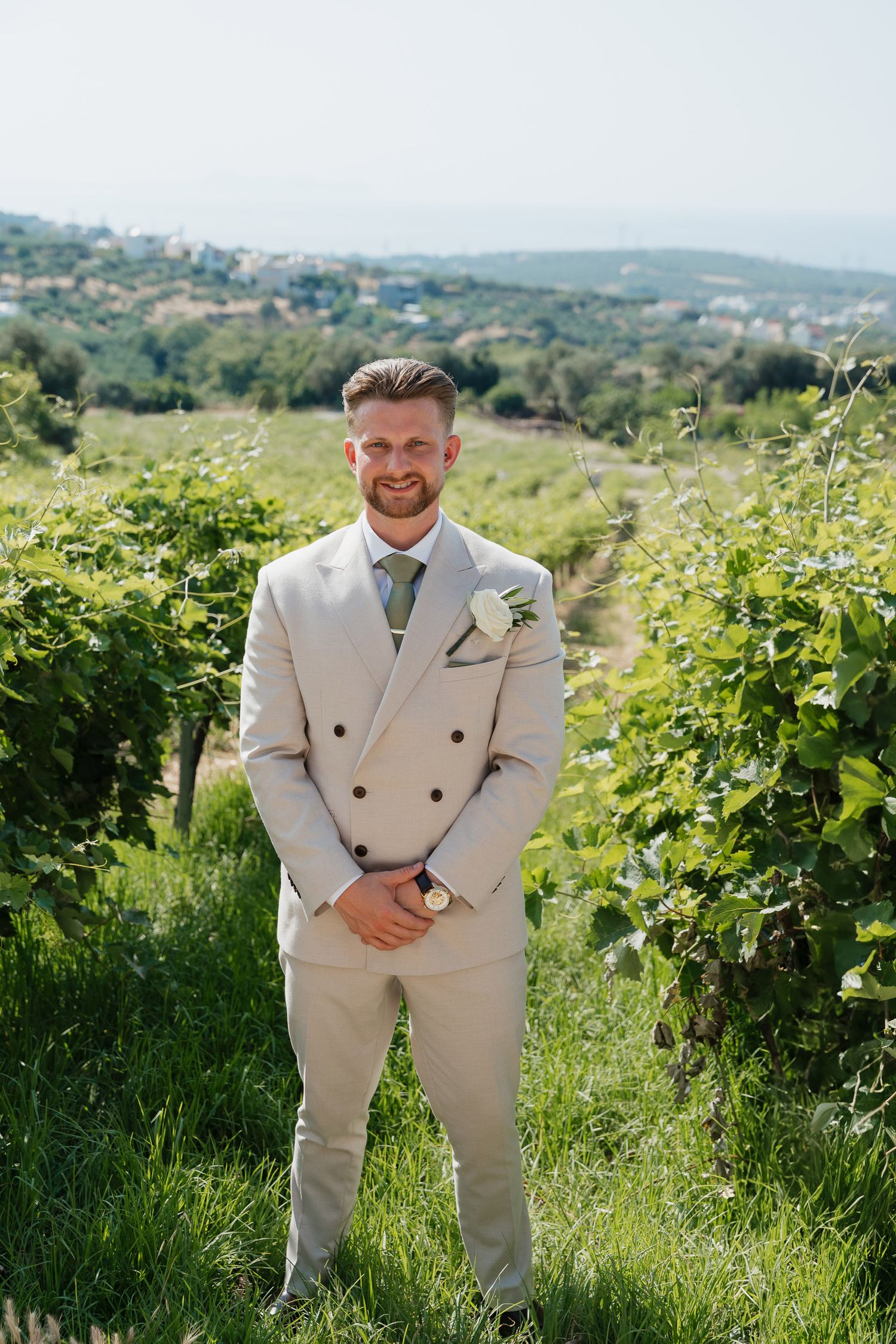 A groom in a cream double-breasted suit stands in a vineyard with rolling hills in the background.