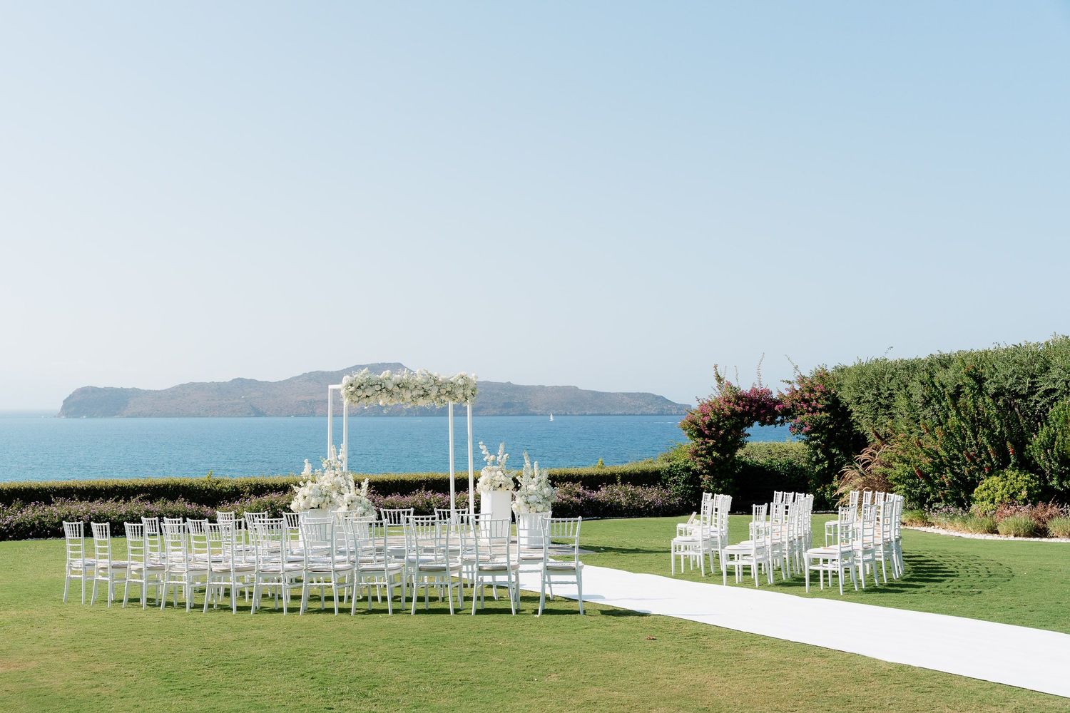 White wedding chairs arranged in rows on a scenic oceanfront lawn with mountains visible in the background.
