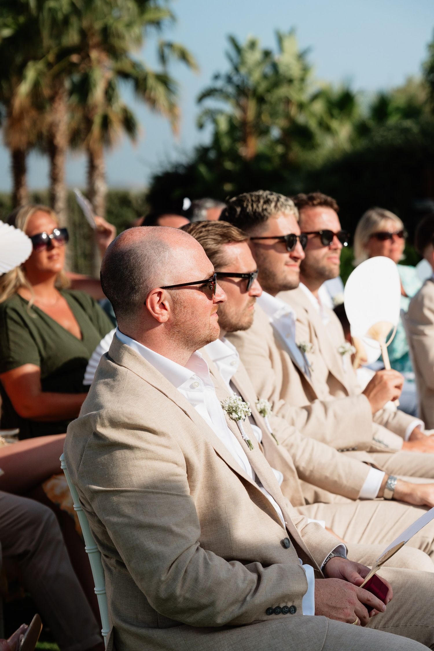 Wedding guests in beige suits and sunglasses sit in a row at an outdoor ceremony with palm trees in the background.