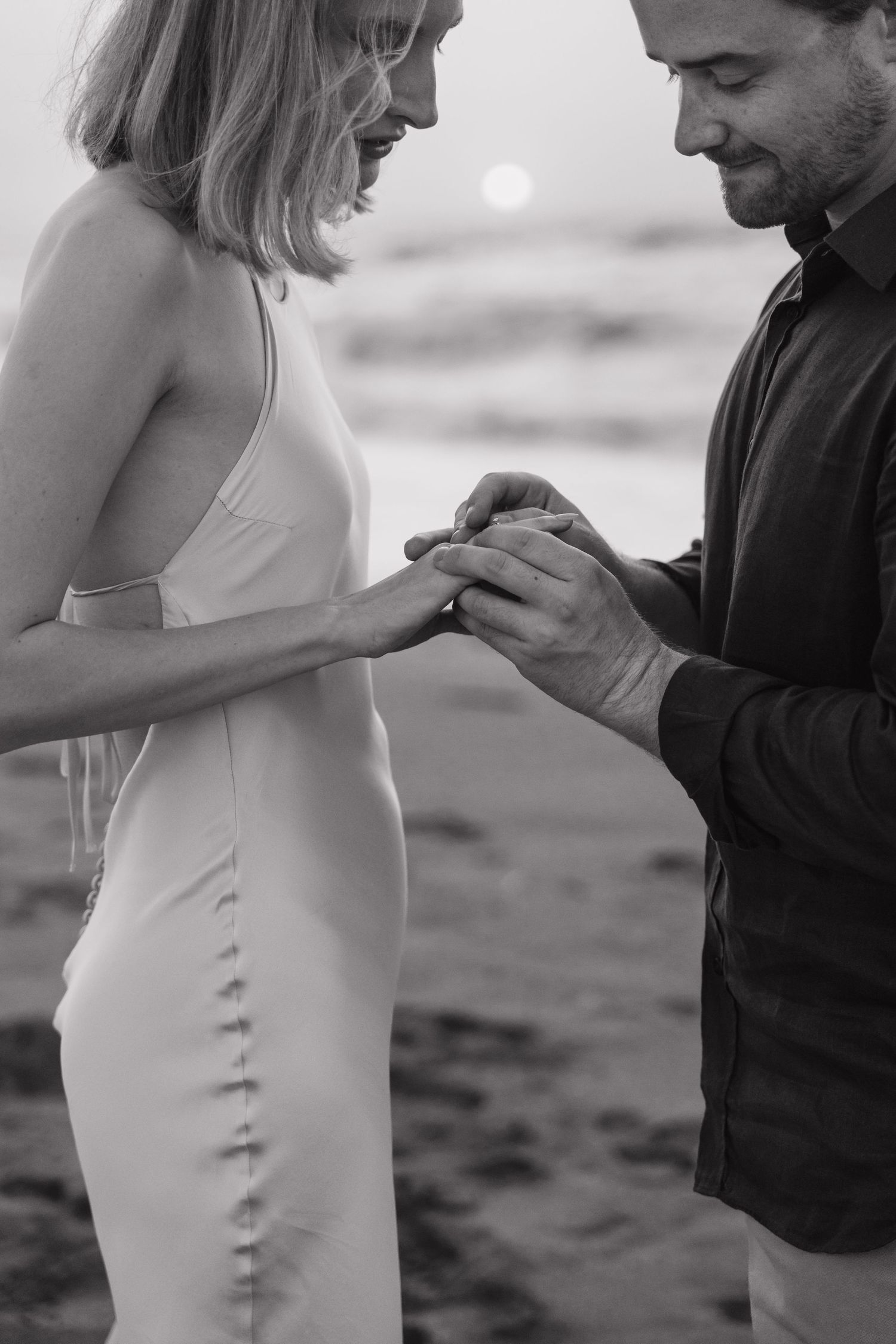 A romantic black and white beach proposal scene with a couple exchanging rings at sunset.