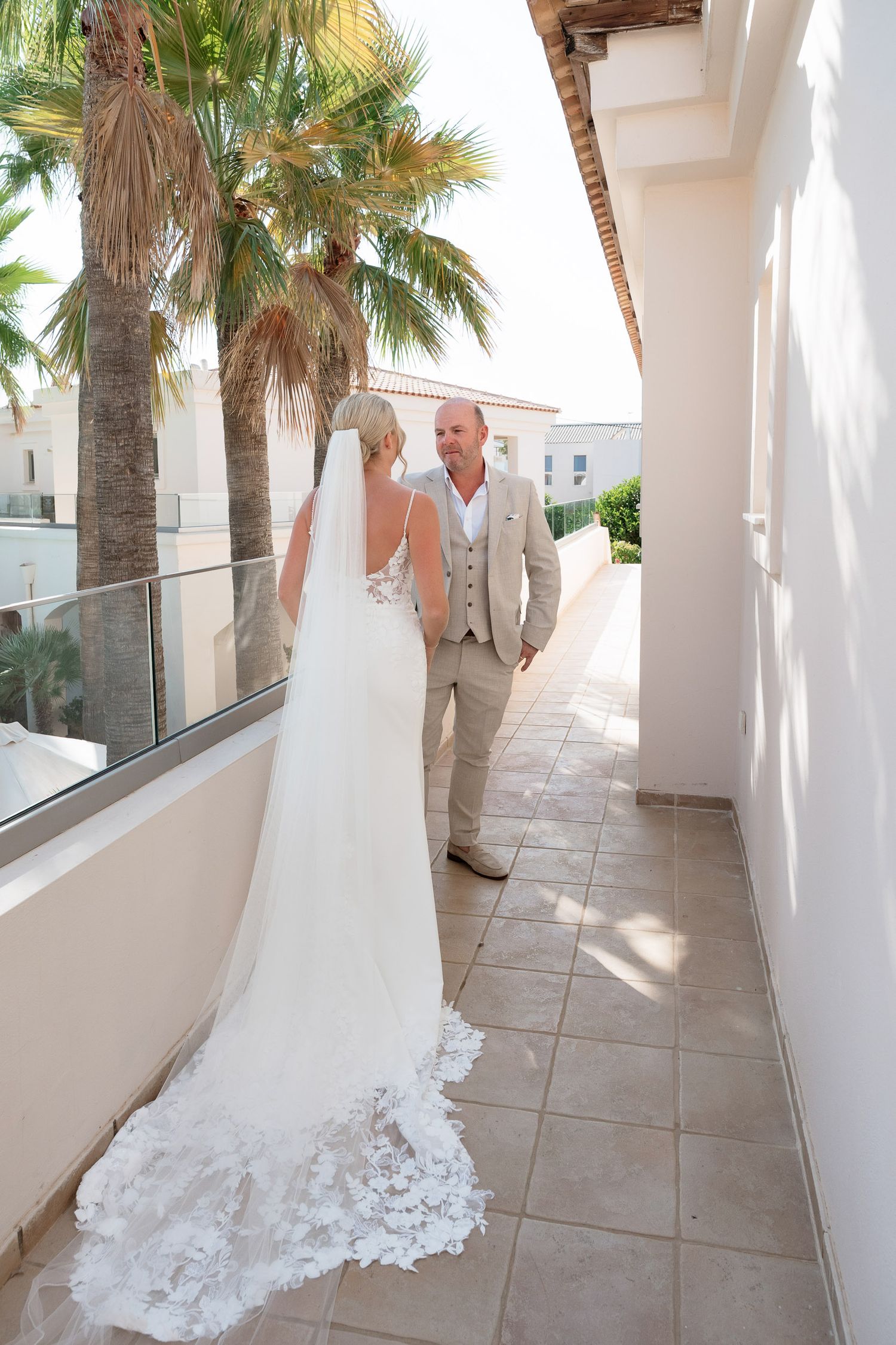 A bright white Mediterranean-style corridor with palm trees and sunlight streaming through arched columns.