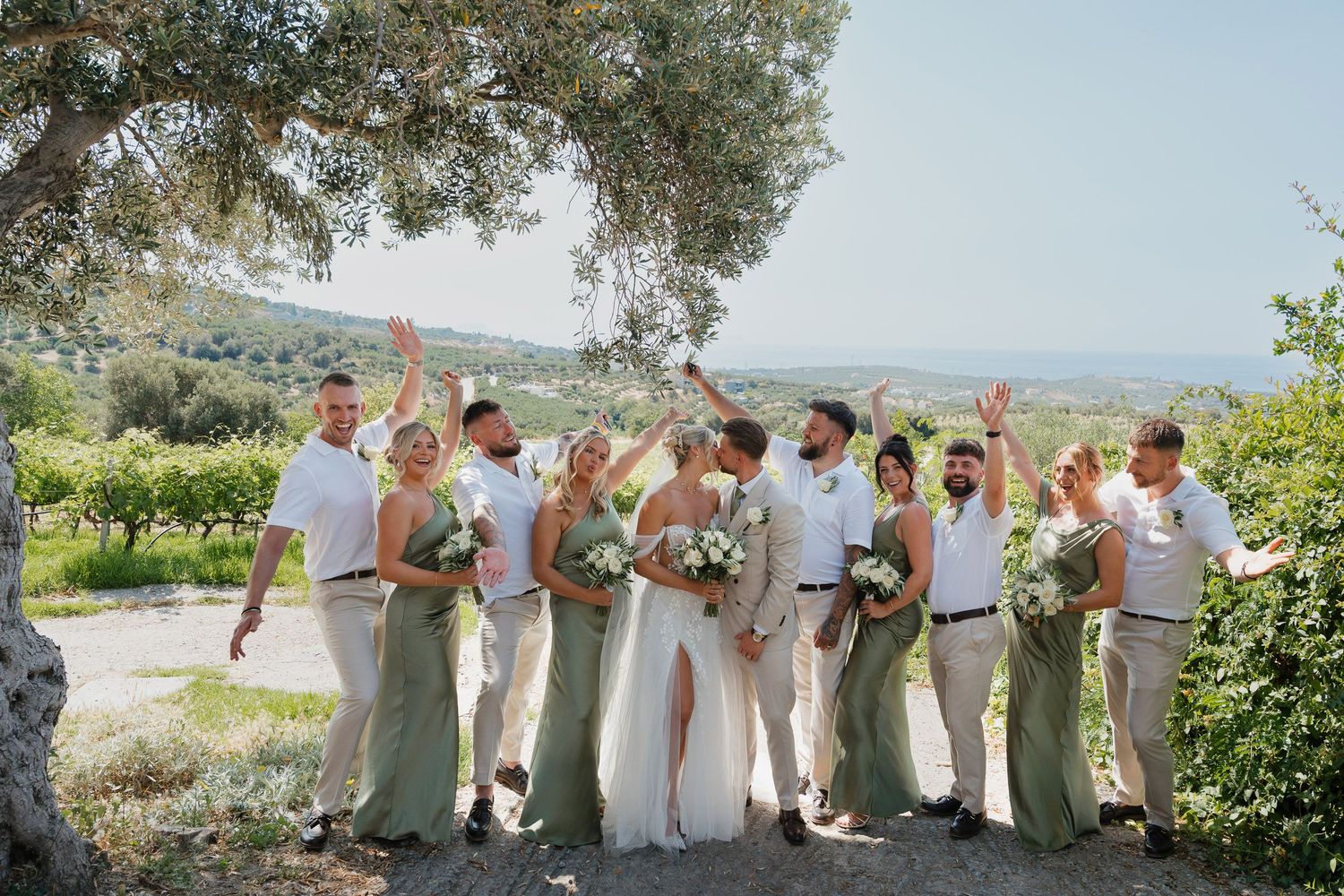Wedding party in light neutral attire poses together on a sunny day with scenic Mediterranean landscape in background.