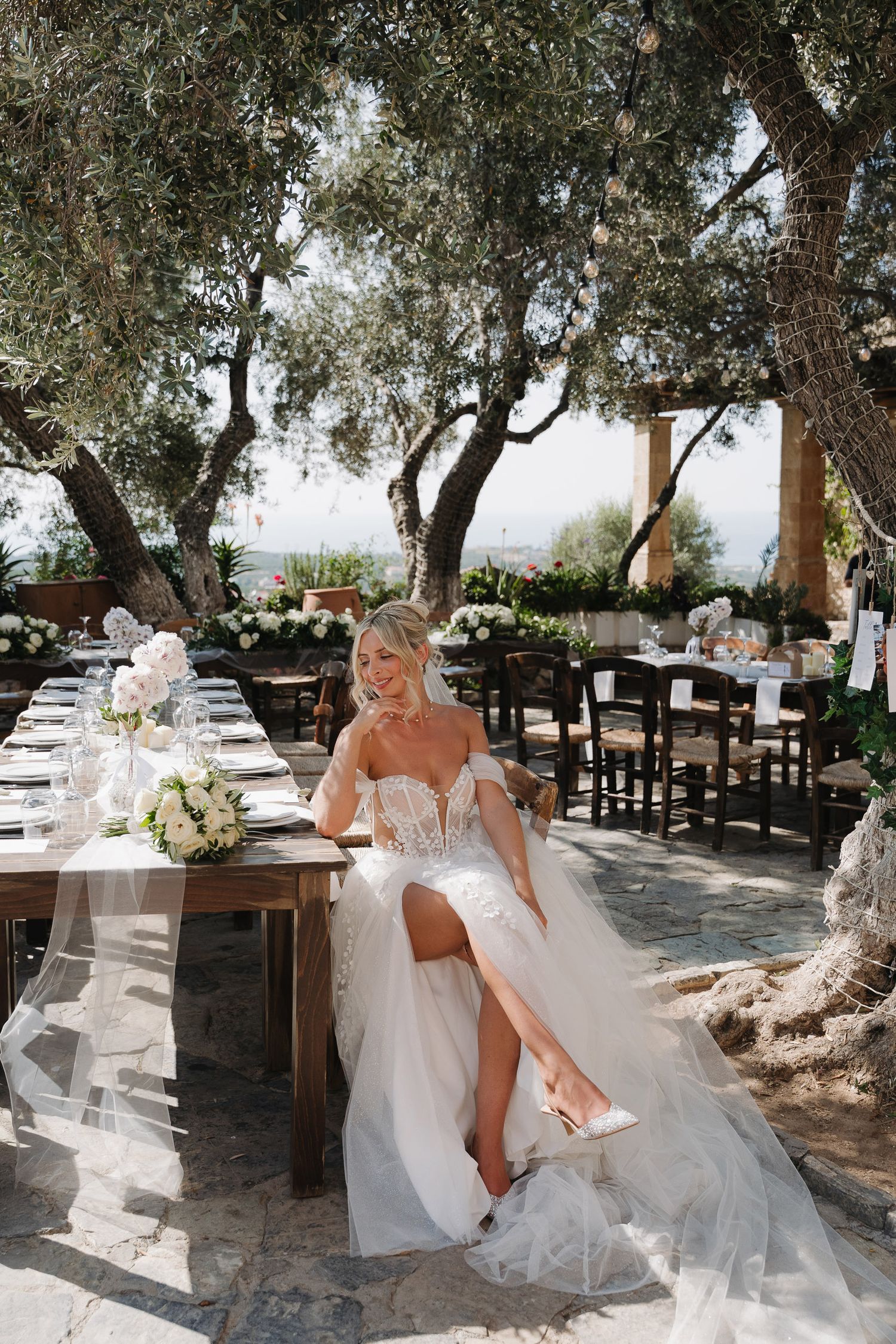 An elegant outdoor wedding reception setup under olive trees with white table settings and floral arrangements on a sunny day.