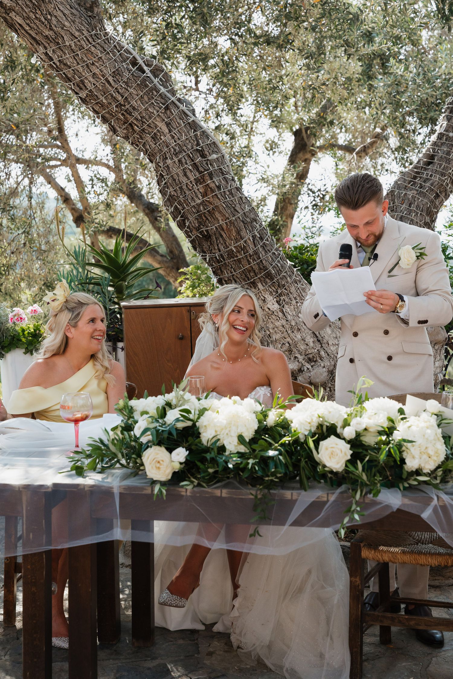 Elegant outdoor wedding reception table decorated with white roses and greenery under a rustic tree canopy.