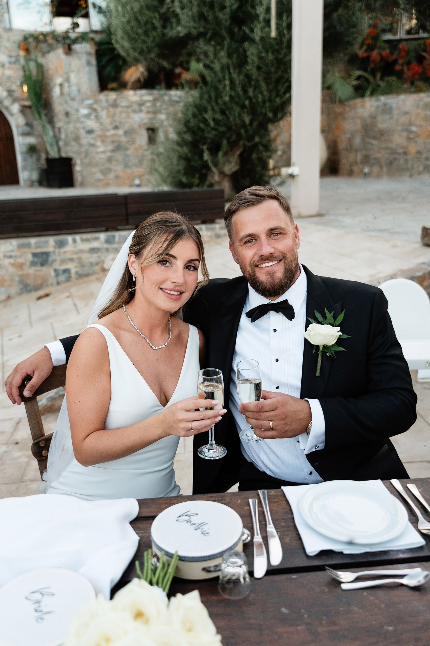 A couple in wedding attire sits at a rustic table sharing drinks and smiles at an outdoor reception.