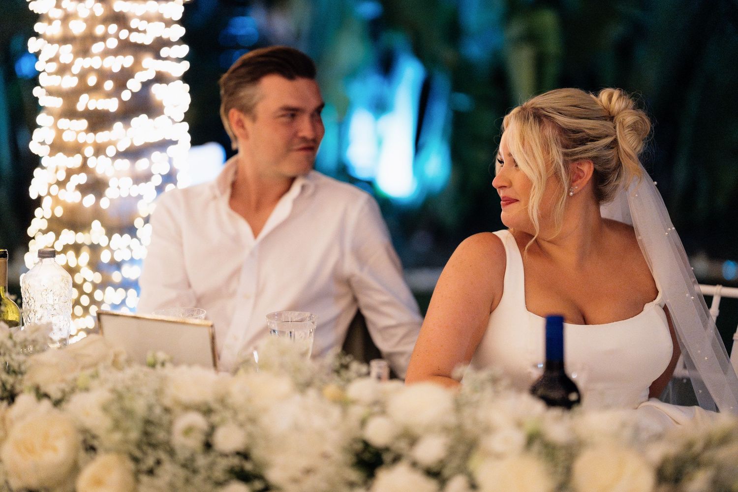 Wedding reception guests seated at an elegant outdoor table decorated with white floral arrangements and twinkling lights.