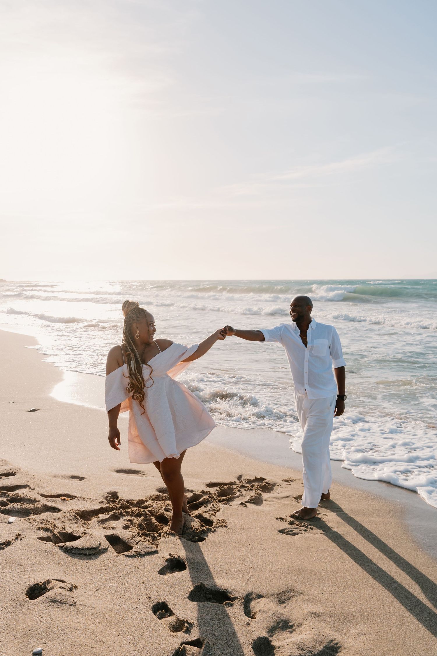 A couple in white clothing walks together along a sandy beach with ocean waves in the background.