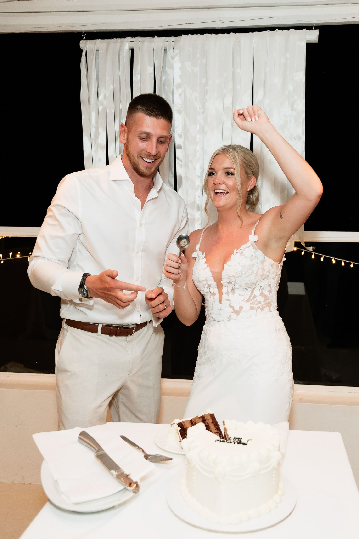 A couple in white attire shares a playful moment while cutting their wedding cake during the reception celebration.
