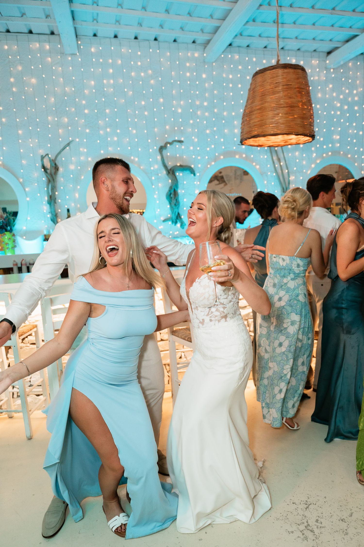 Wedding guests dance and celebrate with joyful energy under turquoise lighting at an indoor reception venue.