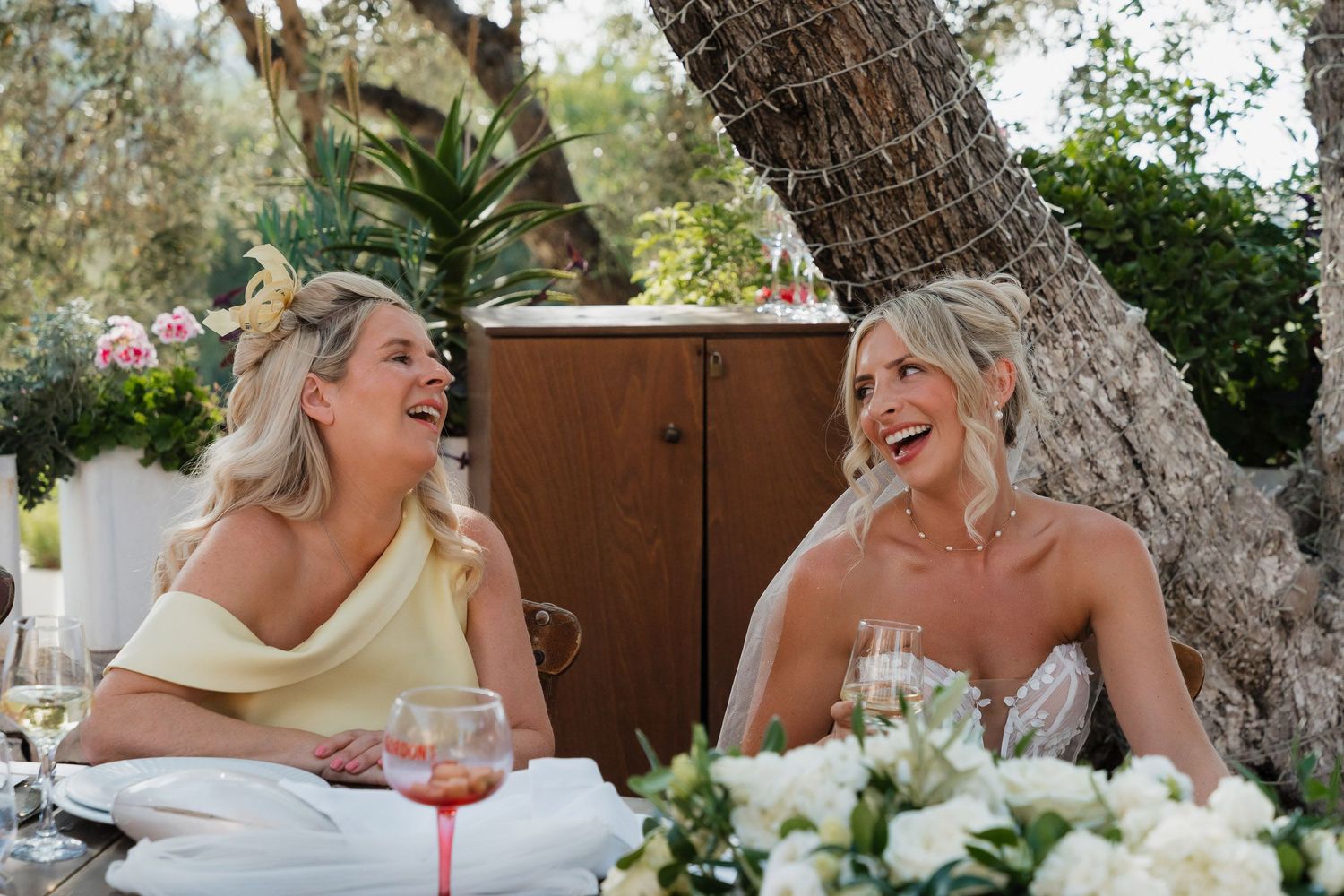 Two people share a joyful laugh while seated at an outdoor table with white floral decorations and wine glasses.