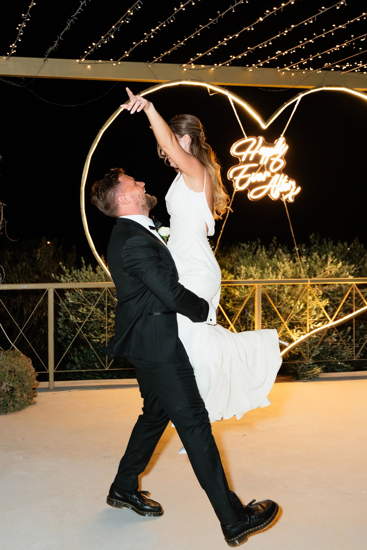 A couple performs an elegant dip during their wedding dance on a white dance floor under glowing lights at night.