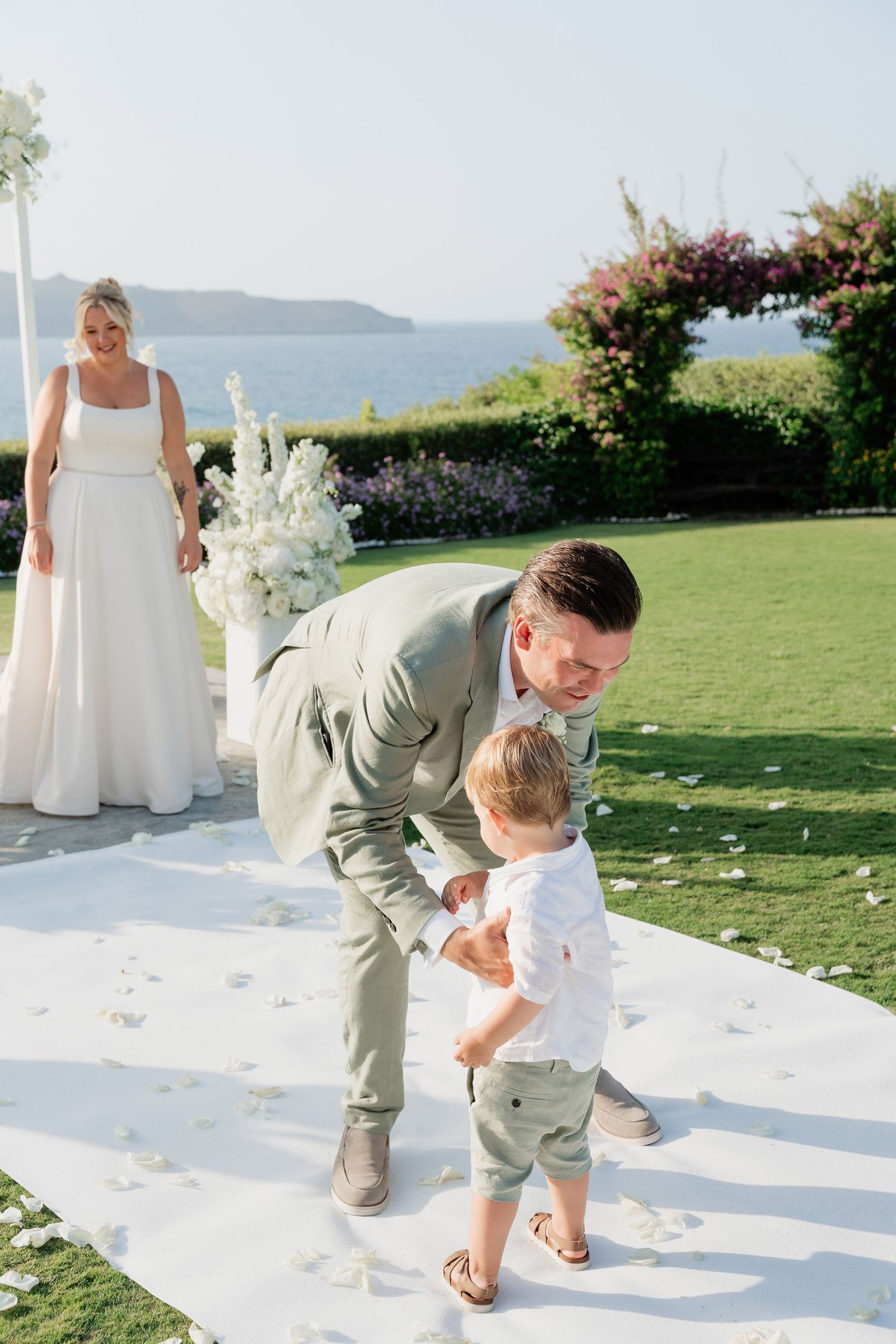 A sweet moment during an outdoor wedding ceremony on a grassy lawn overlooking the ocean with floral decorations.