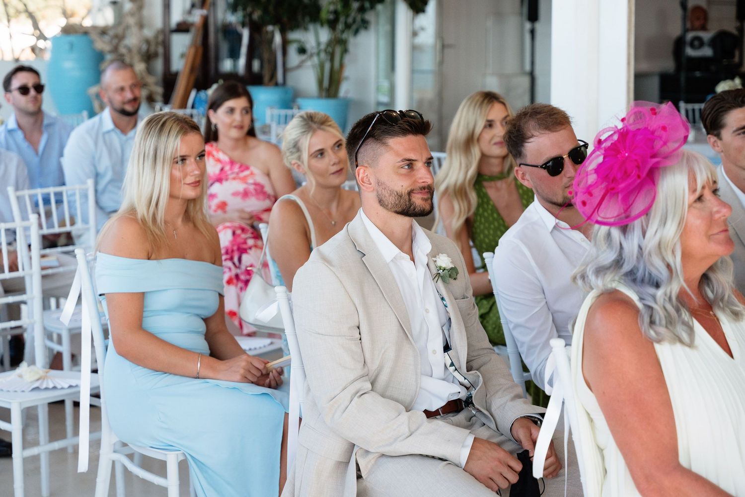 Wedding guests dressed in white attire are seated outdoors at a ceremony with tropical decor and bright pink accents.