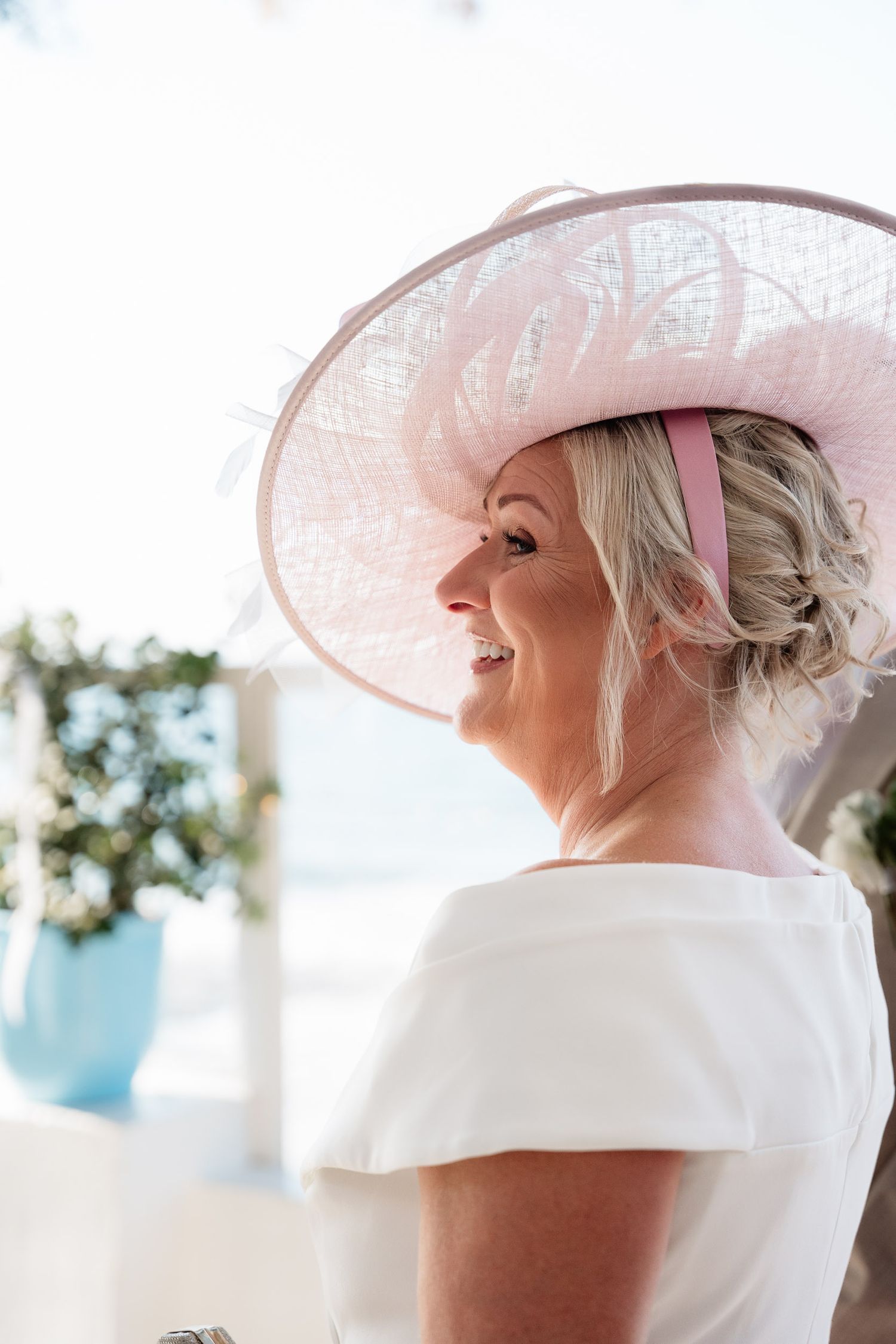 A person wearing an elegant white dress and wide-brimmed pink hat smiles while looking to the side in a bright setting.