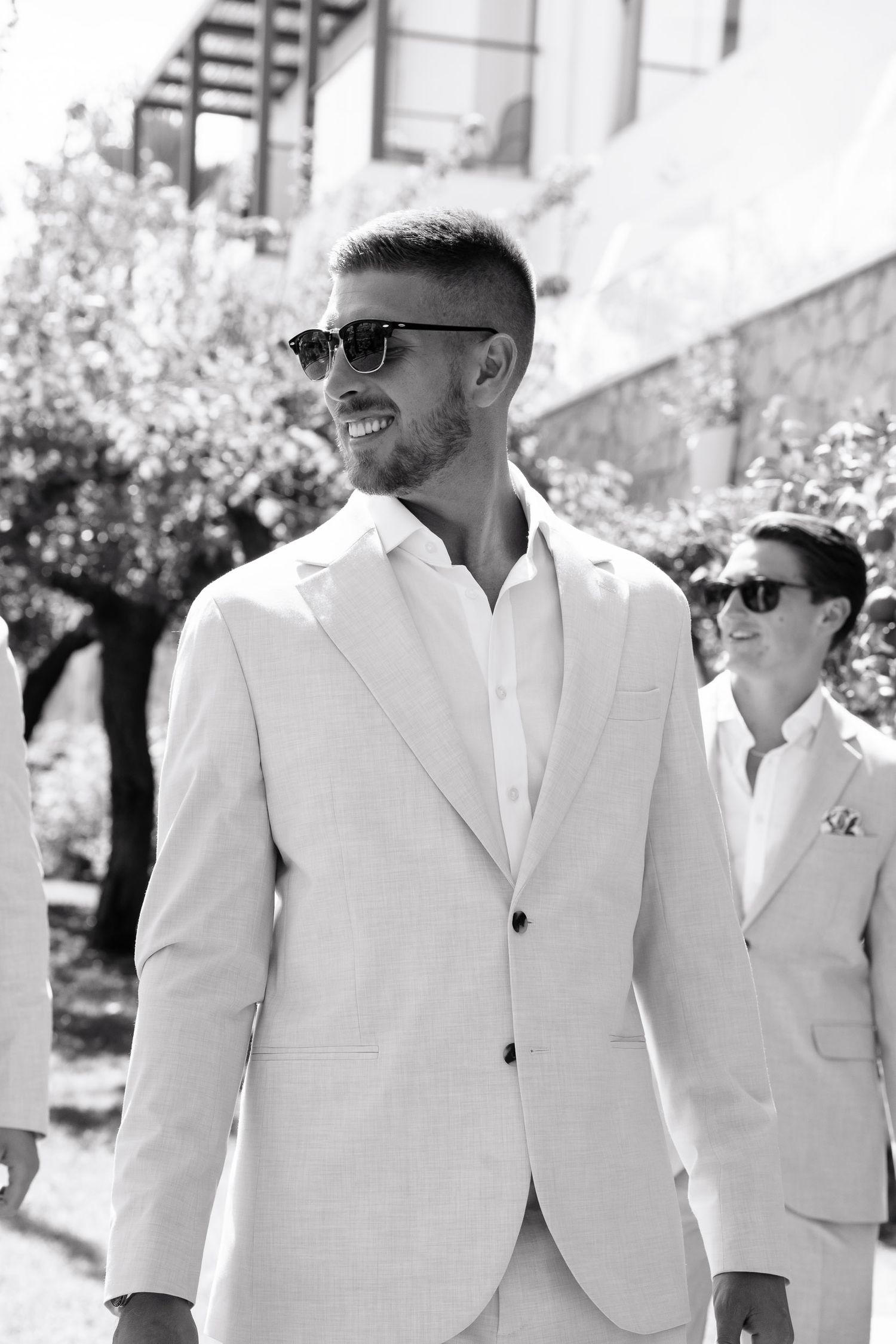 A black and white photo of two people wearing light colored suits and sunglasses at an outdoor event.