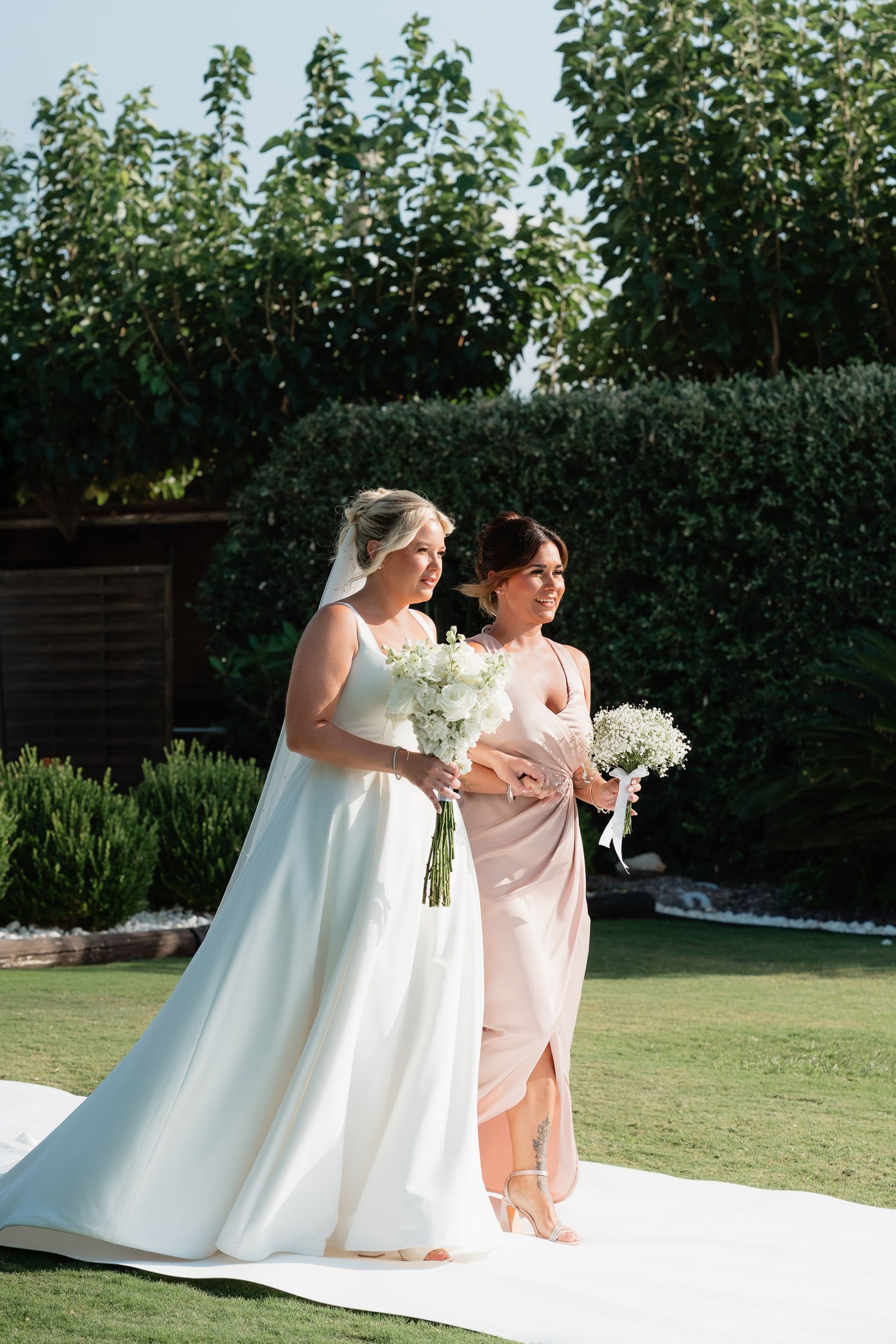 Two people in formal attire share a candid moment of laughter while standing together on a lawn with greenery in background.