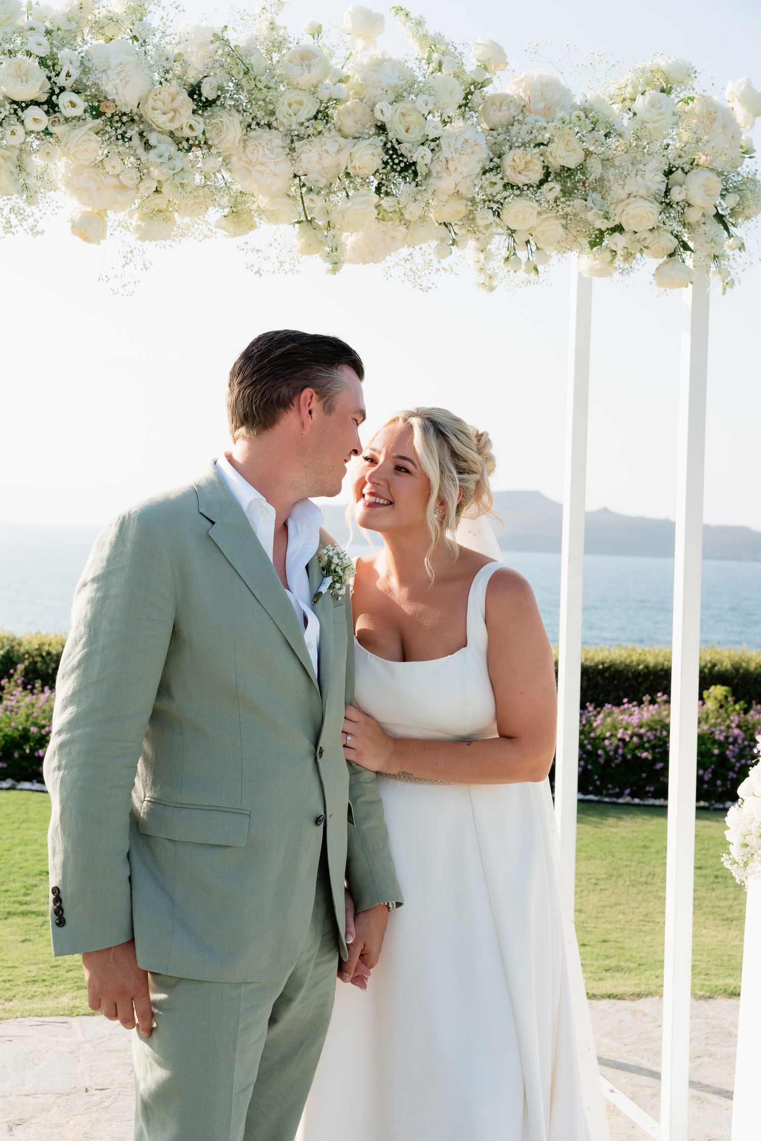 Couple shares romantic moment under white floral wedding arch overlooking ocean on sunny day.