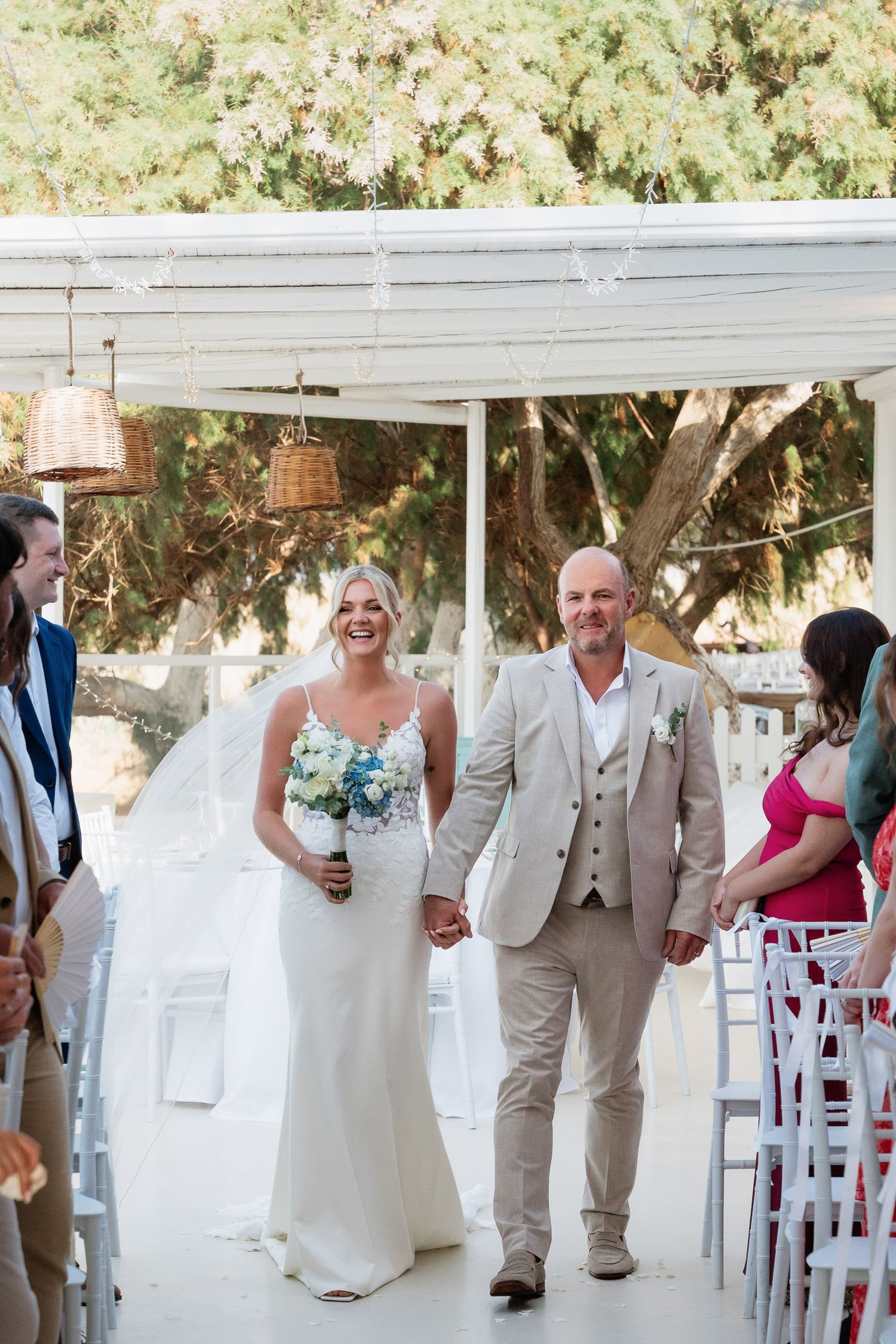 Wedding party walks down outdoor aisle in coordinated white and beige attire at sunny garden ceremony.