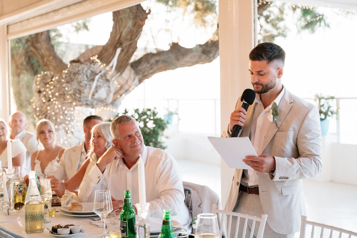 A speaker in a light-colored suit delivers a speech at an elegant outdoor reception with guests seated at tables.