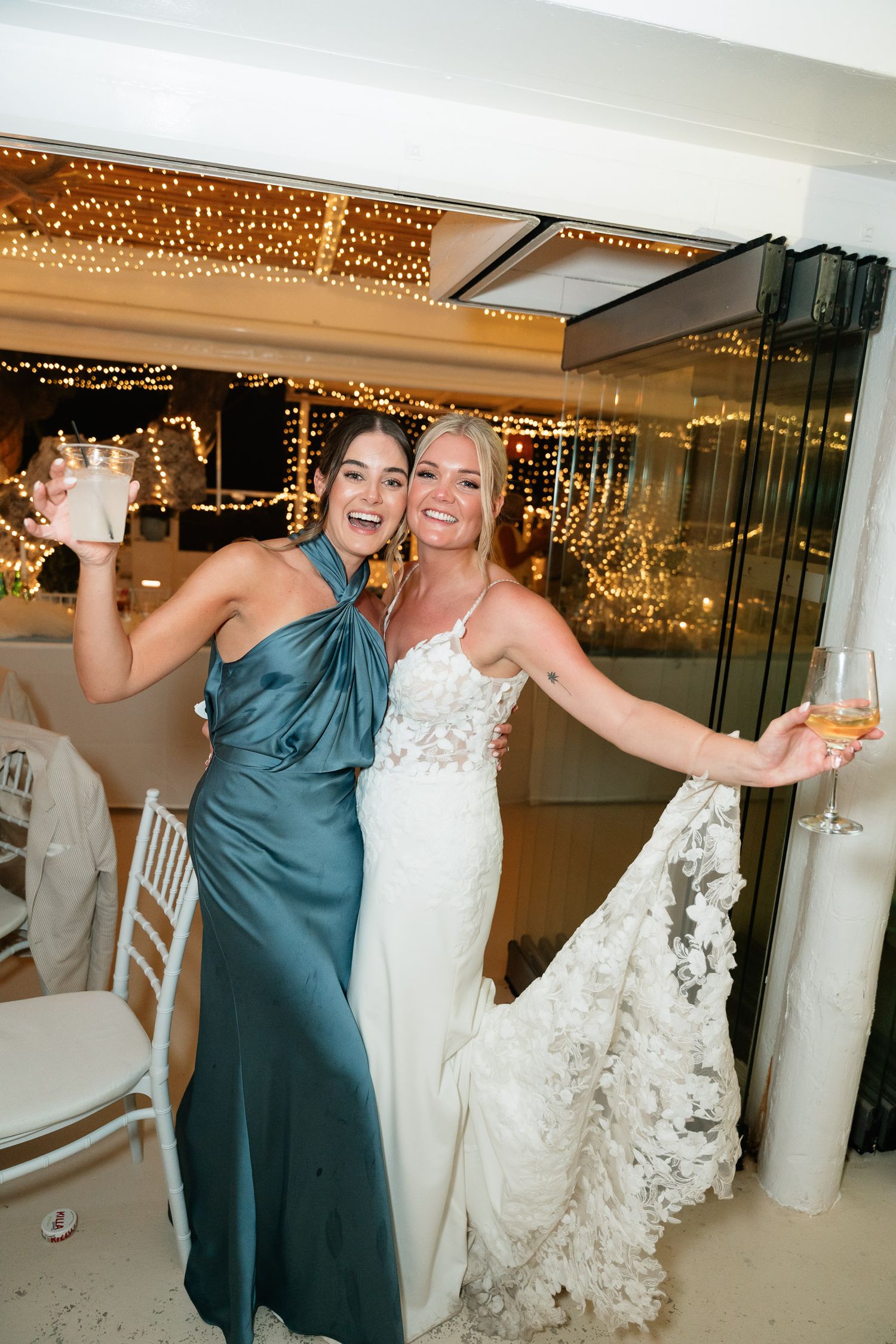 Wedding party members dance and celebrate joyfully under string lights at an indoor reception venue.