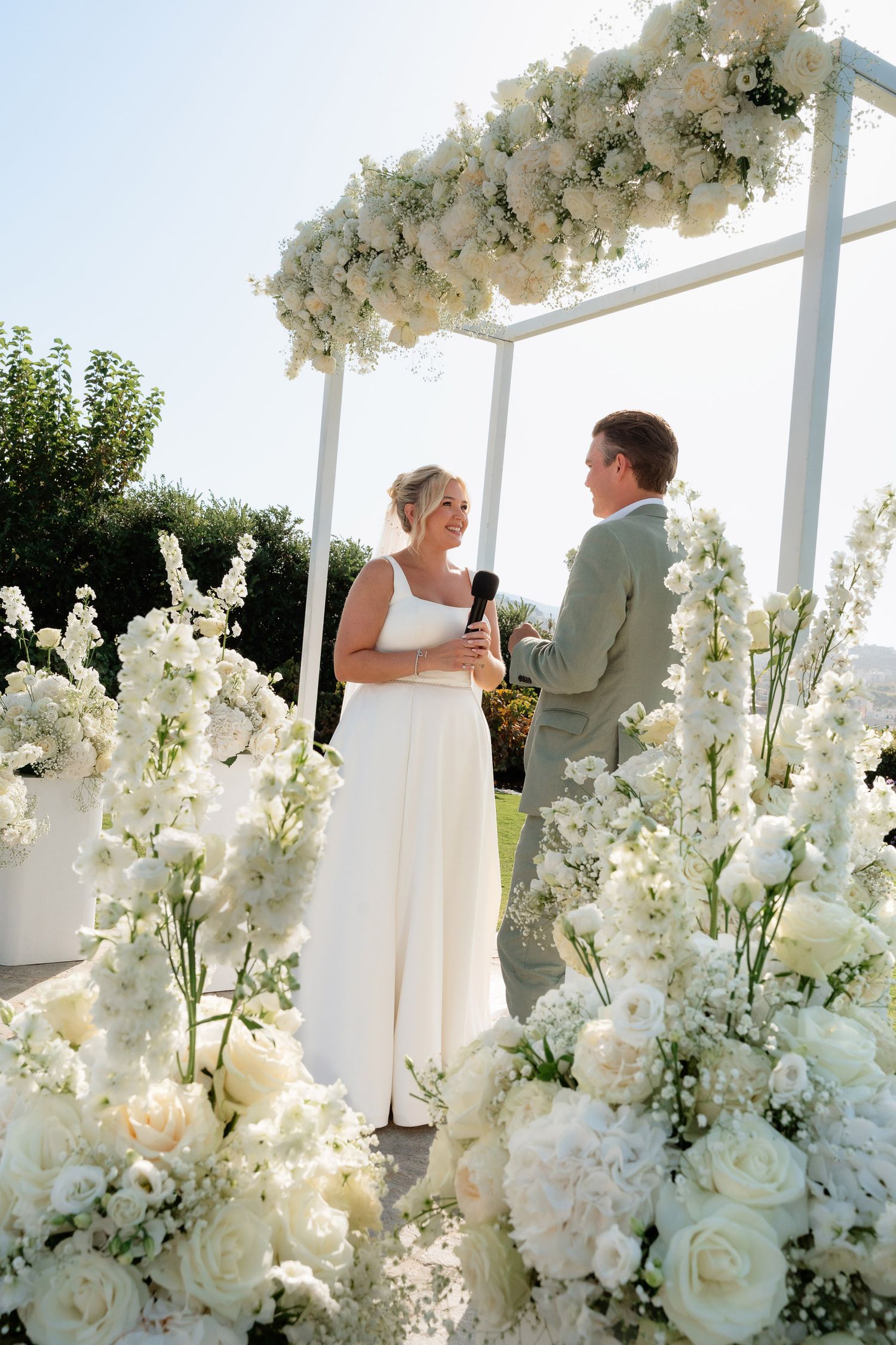 Elegant white floral arrangements with roses and delphinium decorate an outdoor wedding ceremony under a white pergola.