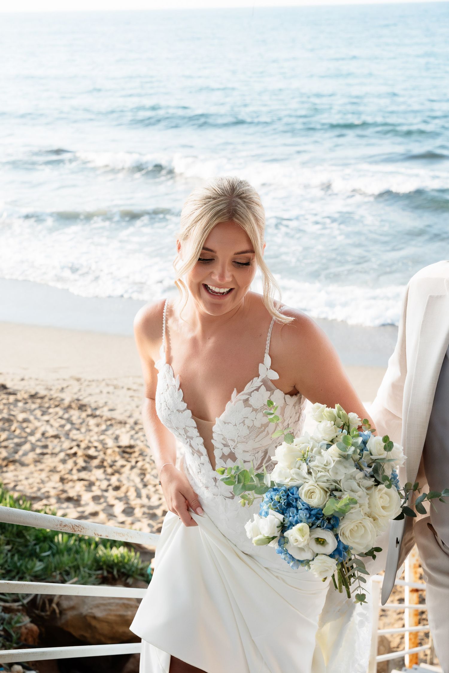 A bride in a white lace wedding dress holds a blue and white floral bouquet while standing on a beach overlook.