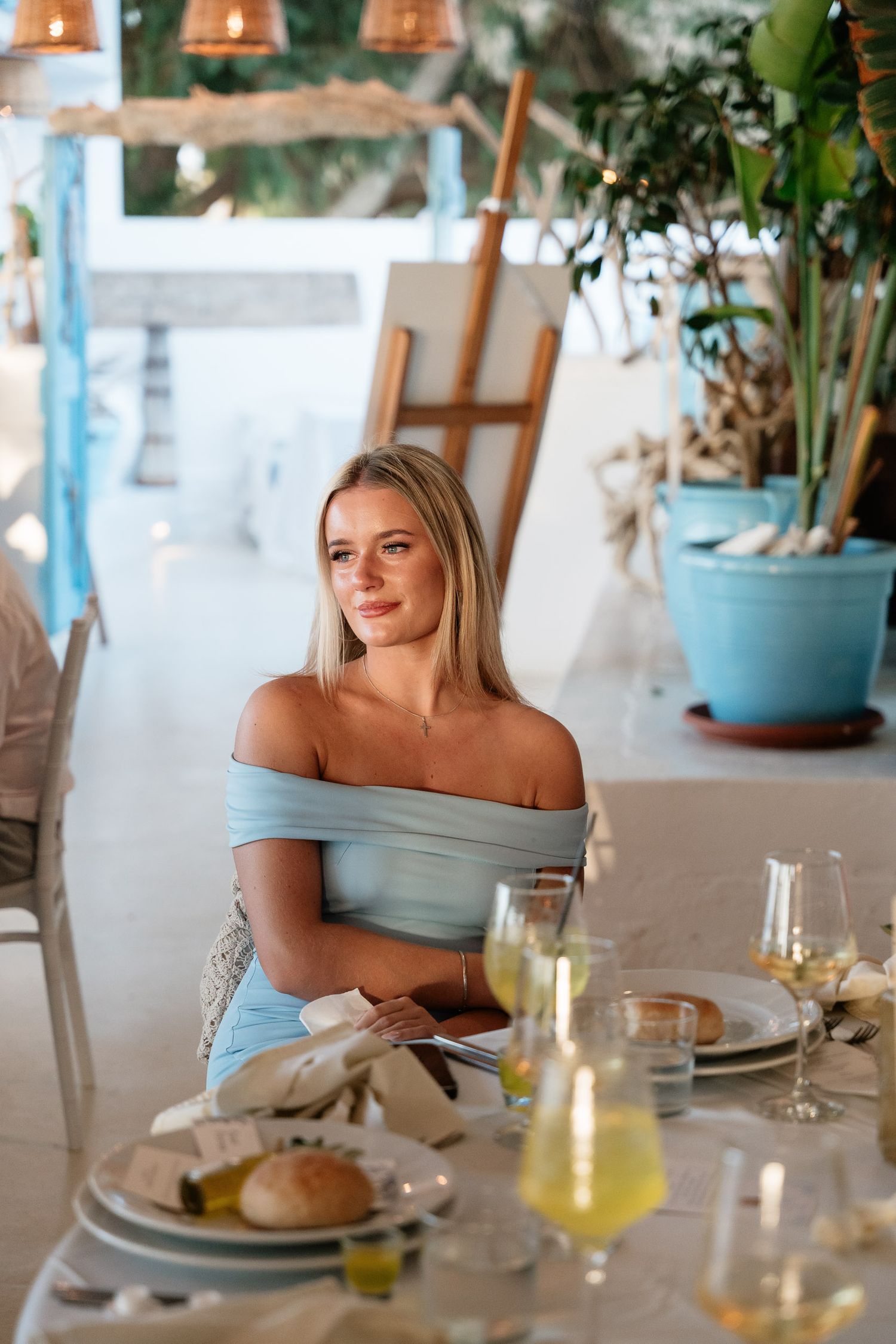 A person in a light blue dress sits at an elegant restaurant table with glassware and place settings.