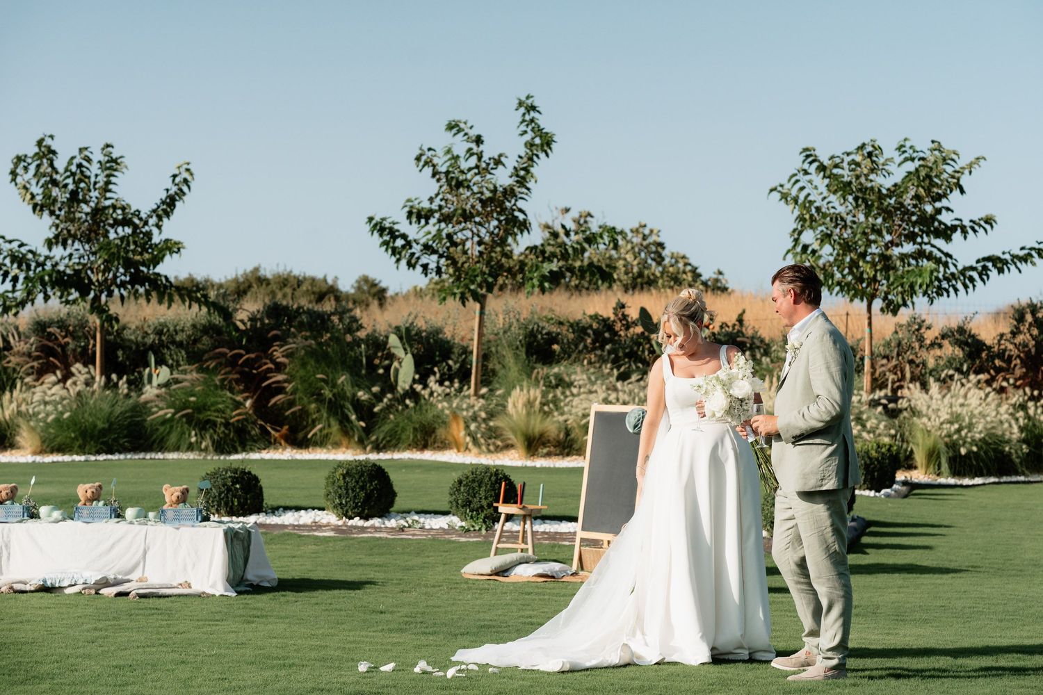 An outdoor wedding ceremony takes place on a manicured lawn with trees and floral arrangements in the background.