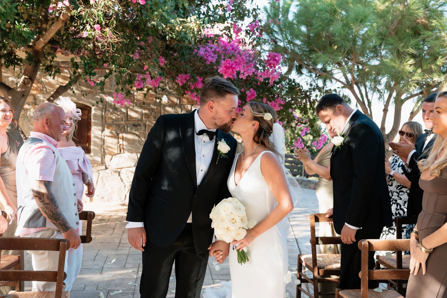 Wedding party celebrates outdoors under trees decorated with pink bougainvillea flowers during a summer ceremony.