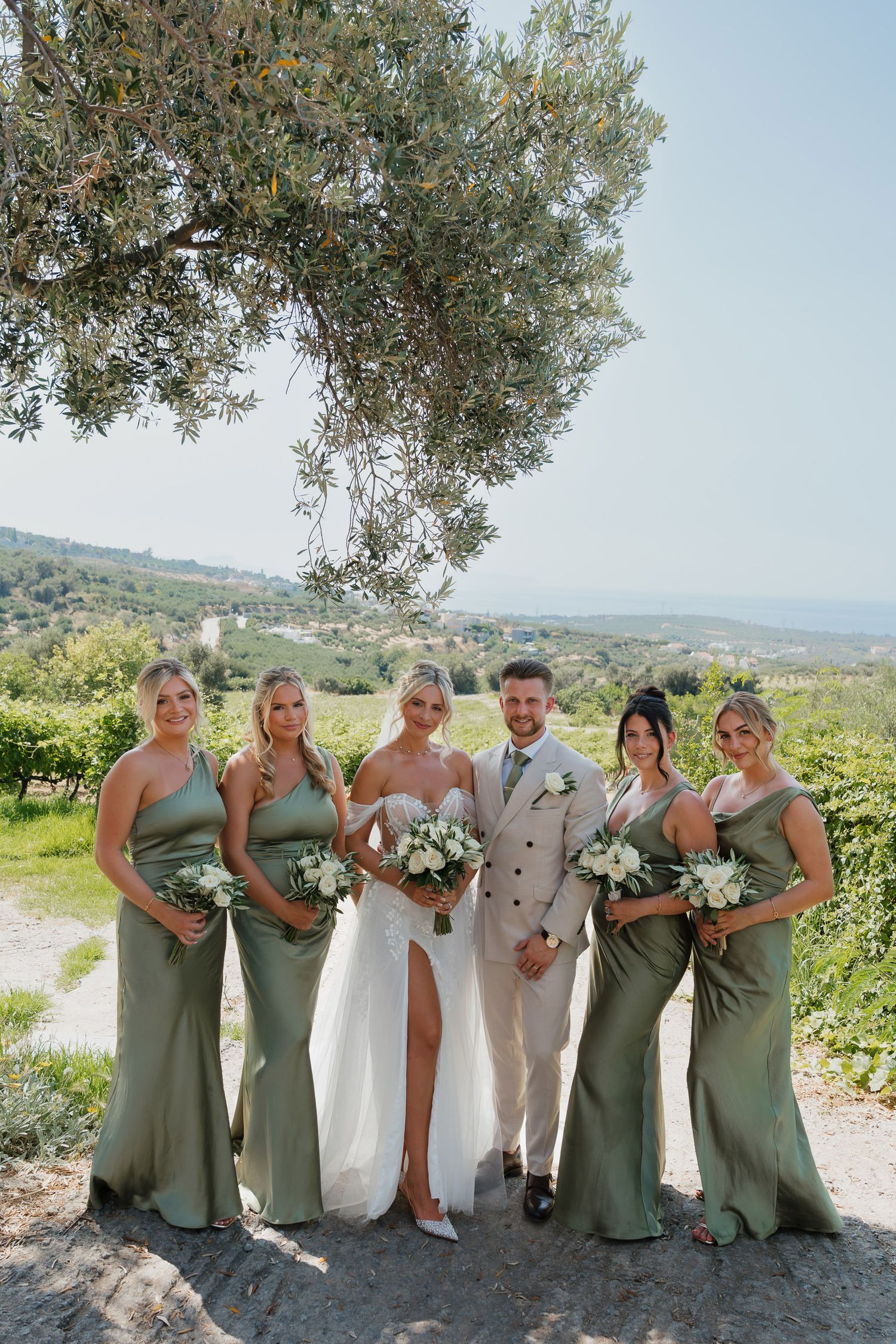 A wedding party poses outdoors in Tuscany with bridesmaids in sage green dresses and an elegant bridal gown against a scenic view.