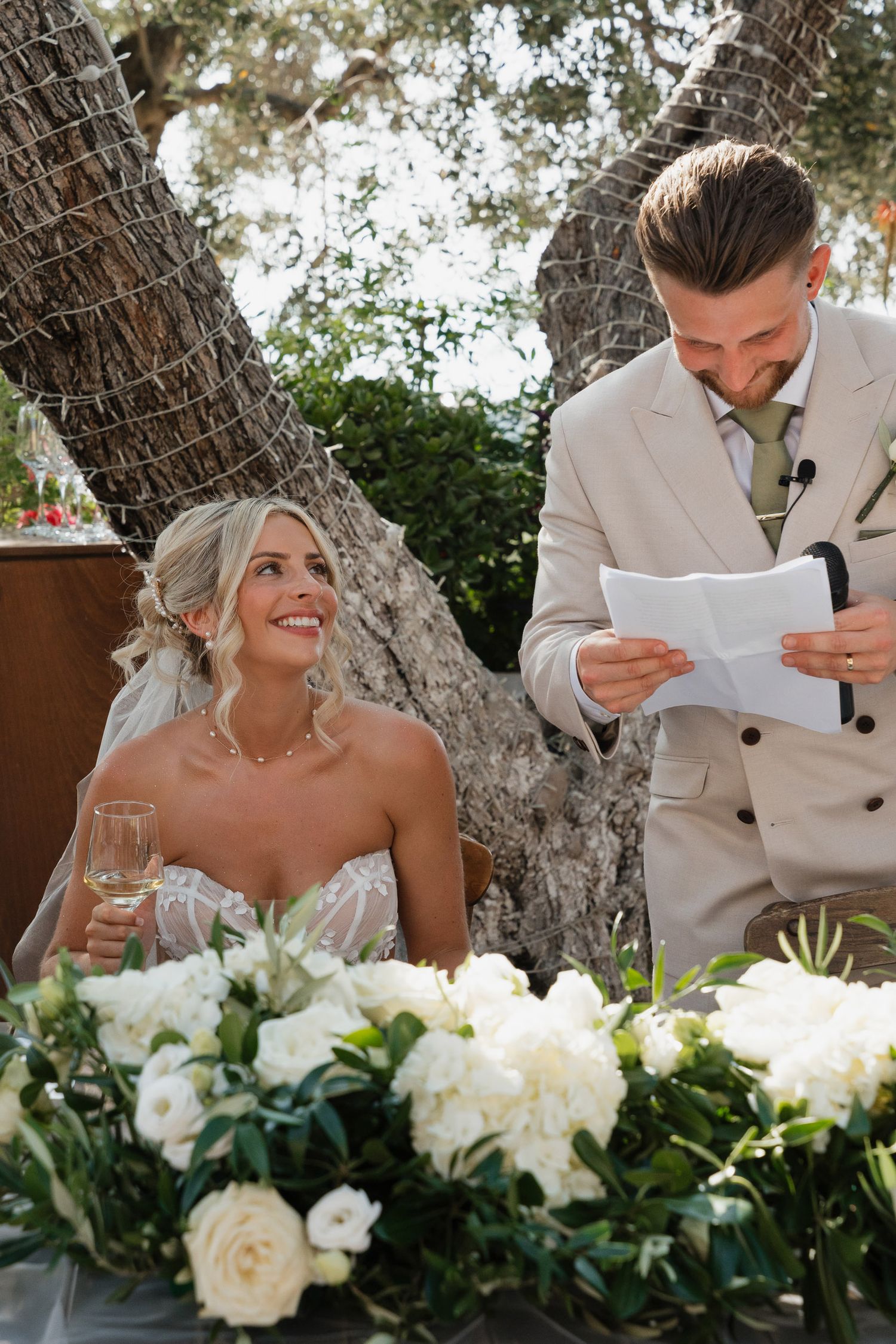 A wedding couple shares a joyful moment during an outdoor ceremony with white floral arrangements and rustic tree backdrop.