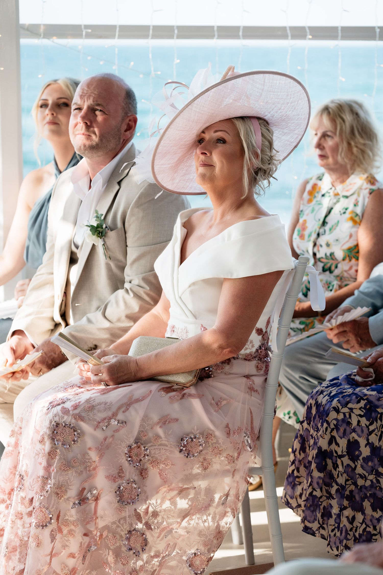 Wedding guests seated in formal attire including a white summer hat and floral dresses at an outdoor ceremony.