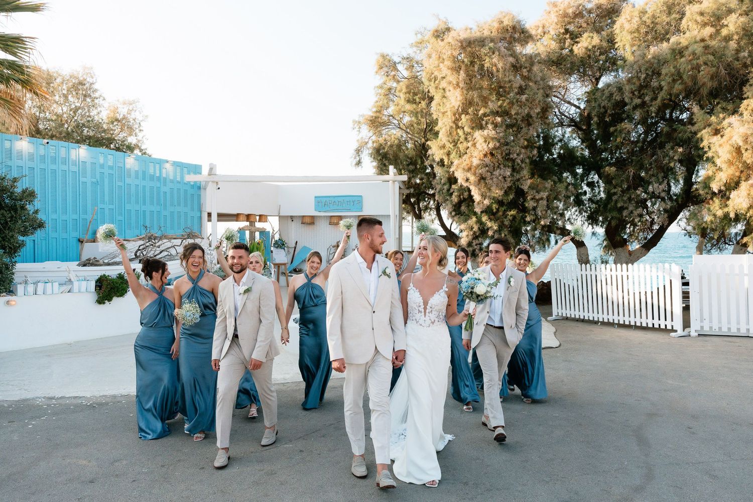 Wedding party in teal blue dresses and light gray suits walk together on a white picket fence lined path at sunset.