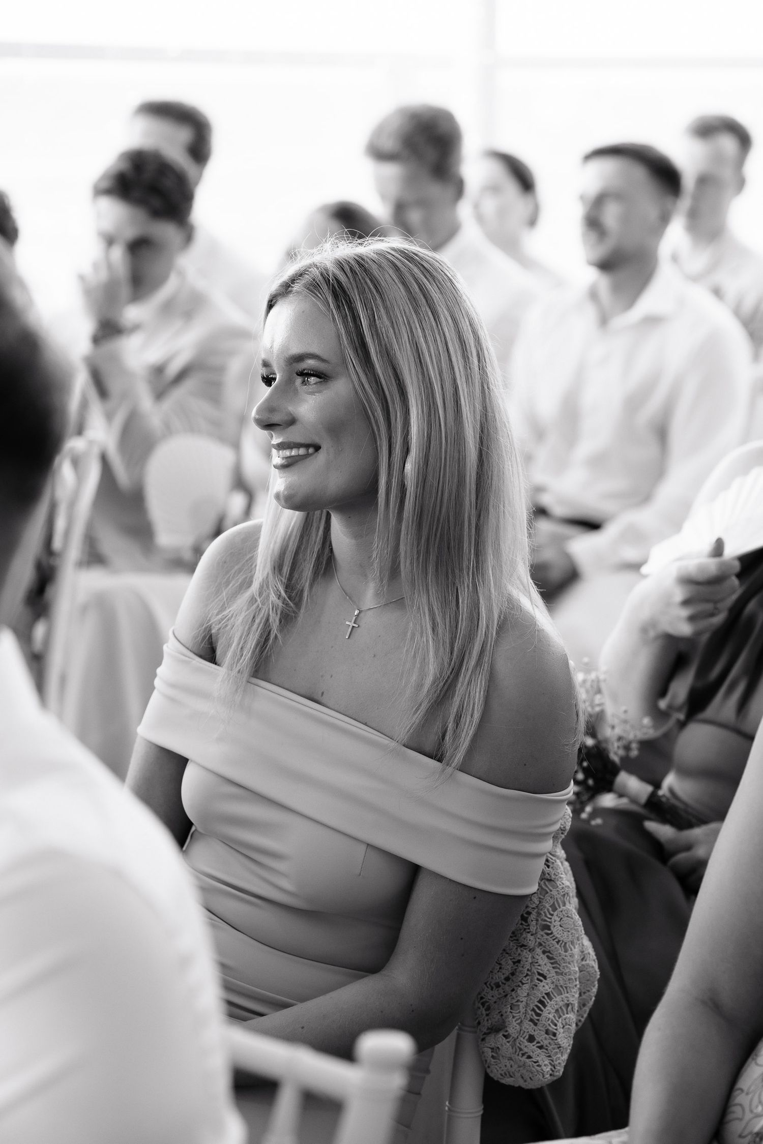 A candid black and white photo of someone in an off-shoulder top smiling at an event with people in the background.