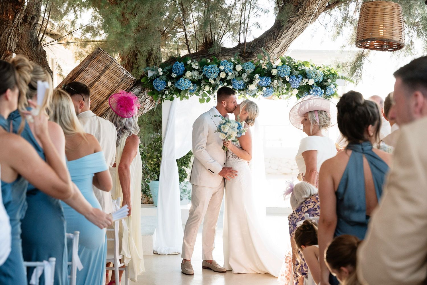 A beautiful wedding ceremony under a floral arch with blue flowers and guests in summer attire.